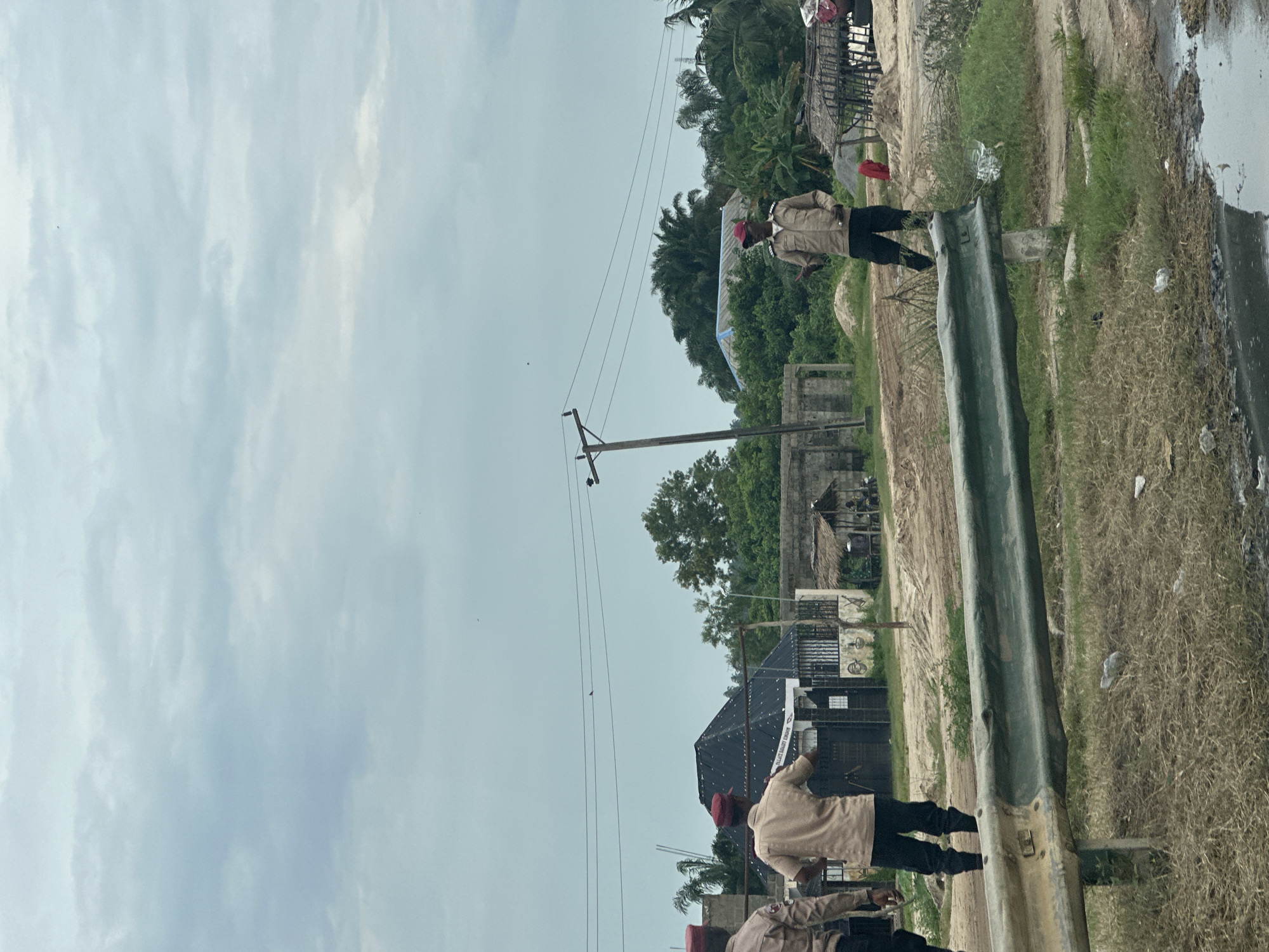 The image depicts a street scene in Badagry, Nigeria. Three individuals, dressed in tan uniforms with maroon caps, stand along a roadside. The foreground features a weathered green metal guardrail and overgrown grass with scattered debris, including a puddle of dark water. In the background, there are various structures, including a house with a dark, pitched roof, fences, and a thatched-roof hut. Electrical poles and power lines stretch across the frame. The overall environment appears to be a mix of residential and possibly undeveloped land, under a cloudy sky.