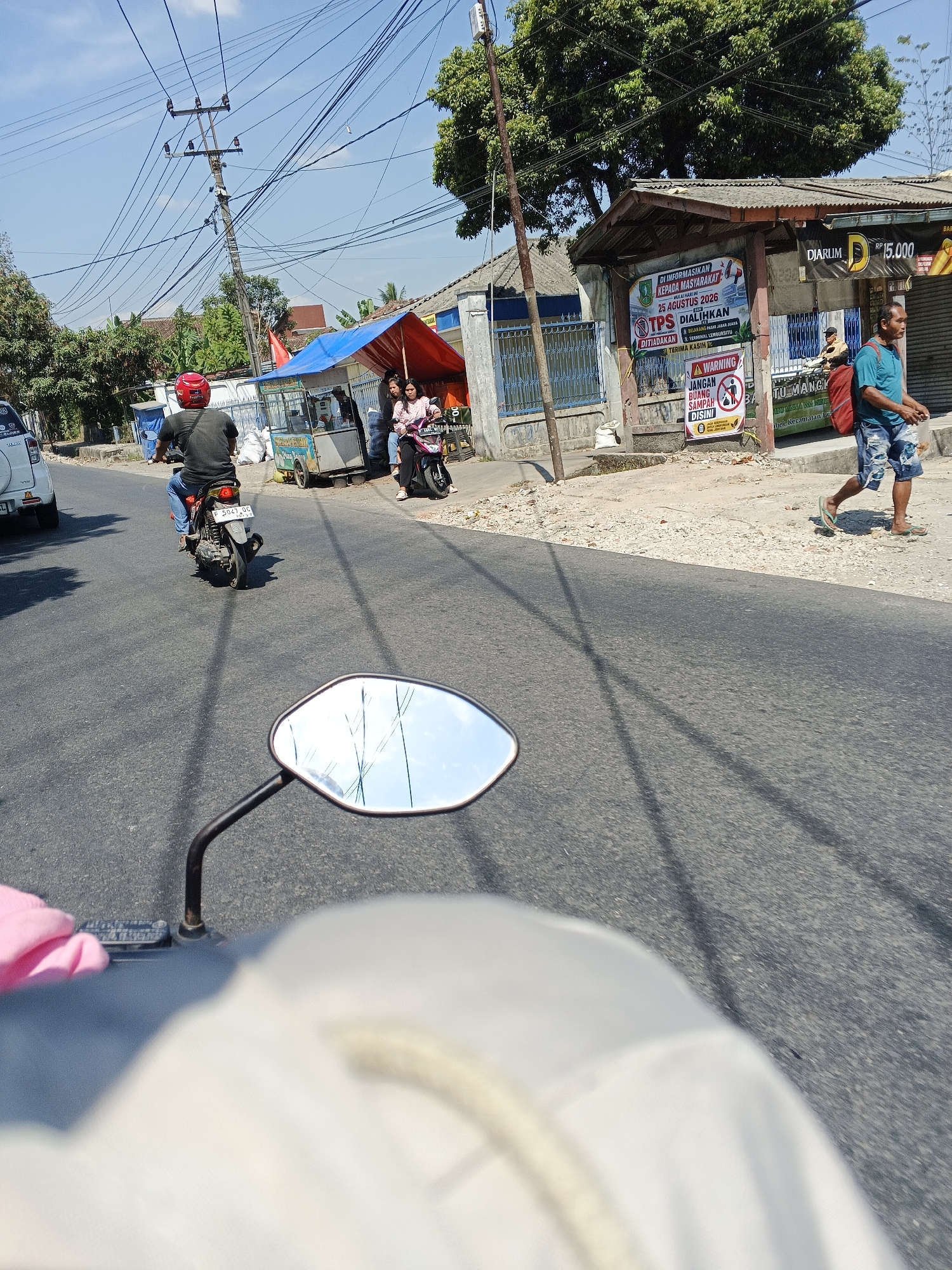 In the city of Sukabumi, Indonesia, on a bright, sunny day, the image captures a street scene. The perspective is from the viewpoint of someone riding a motorcycle, indicated by the motorcycle's mirror and part of the handlebar in the foreground. The road is paved with asphalt, and power lines crisscross the sky above.
On the right side of the road, there are several structures. One appears to be a small shop or stall with a corrugated metal roof. A sign on this structure indicates "Diarum Rp 15.000". Next to it, there is another building with a similar roof and blue fencing. A prominent poster on the wall of this building announces "DIINFORMASIKAN KEPADA MASYARAKAT TPS DIALIKHAN 25 AGUSTUS 2026" and "DILARANG BUANG SAMPAH DISINI" which translates to "INFORMATION TO THE PUBLIC TPS RELOCATED AUGUST 25, 2026" and "DO NOT DUMP TRASH HERE".
Several people are visible. A man in a blue t-shirt and denim shorts walks on the sidewalk, carrying a red bag over his shoulder. In front of the shops, two women are on a pink motorcycle. Further down the road, a man wearing a red helmet is riding a motorcycle, with his back to the camera, and is dressed in a dark shirt and blue jeans. A white car is also visible on the left side of the road. The environment appears to be a somewhat commercial or residential area with trees and buildings lining the street. The sky is clear and blue.