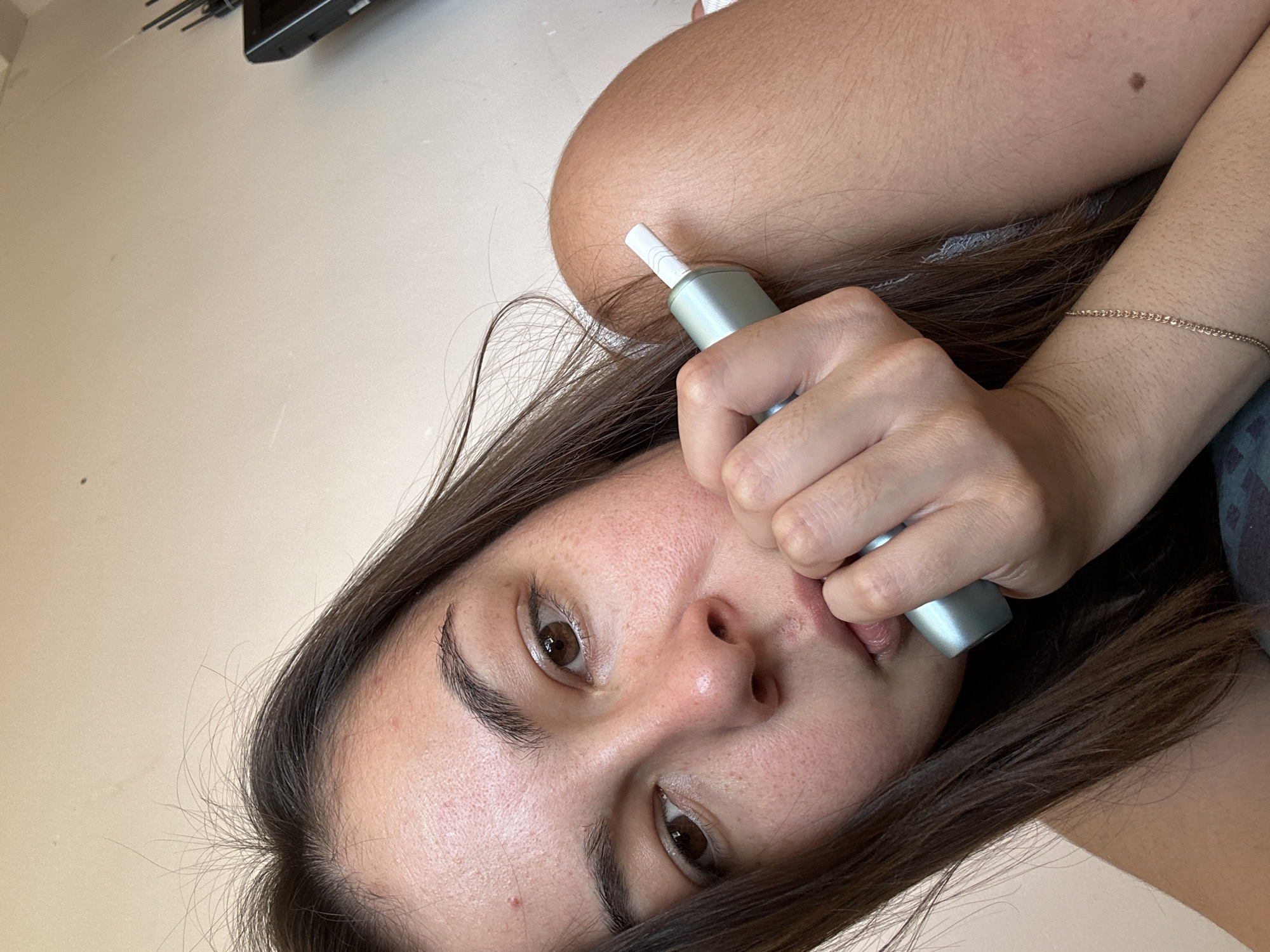 A young woman with brown hair and brown eyes is holding a light blue electronic cigarette device to her lips. She is lying down, with her head tilted to the side. Her right arm is bent, and her hand is resting on her chin. The background is a plain, light-colored wall. There is a black electronic device with antennas visible in the upper right corner of the image. The woman is wearing a simple gold chain bracelet. The image is a close-up, focusing on the woman's face and the device. There is no indication of the location being in Gyumri, Armenia.