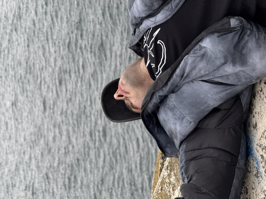 A male individual is depicted lying on his back on a concrete structure, likely a pier or breakwater, along the Black Sea coast in Varna, Bulgaria. He wears a black baseball cap, a black t-shirt with a white abstract design, and a grey and black padded jacket, partially unzipped. His head is tilted upwards, and his eyes appear closed or mostly closed, indicating a state of rest. The concrete surface beneath him shows some faint blue markings. The background consists of the grey, rippled surface of the Black Sea, extending to an unidentifiable horizon under an overcast sky.