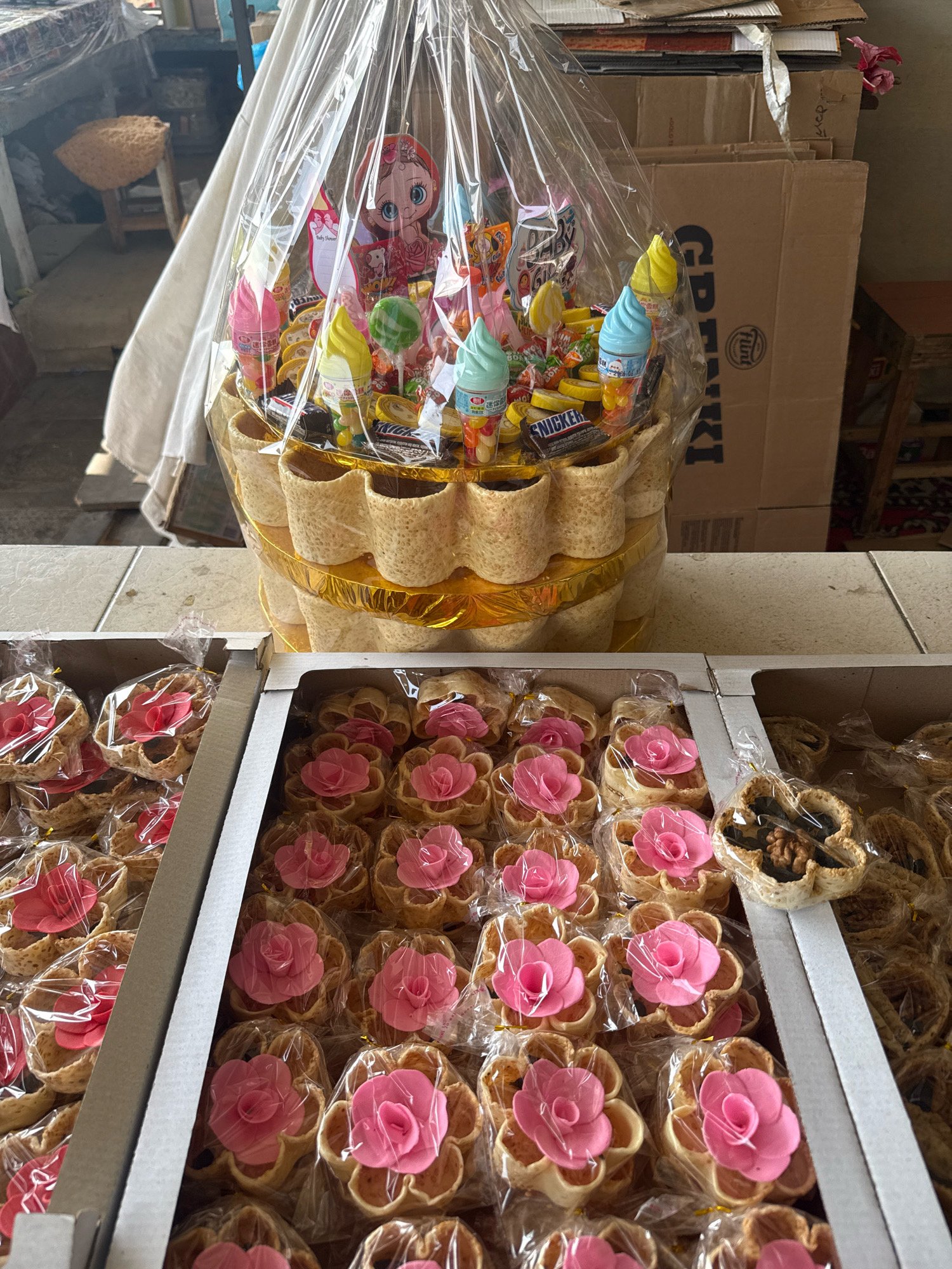 A gift basket filled with candy and snacks is displayed on a tiled surface in an outdoor market stall. The basket is wrapped in clear plastic and features an assortment of colorful candies, including ice cream-shaped lollipops and chocolate bars. In front of the basket, two cardboard boxes are filled with individually wrapped pastries, each adorned with a pink floral decoration. The background shows stacks of cardboard boxes, a wooden stool, and a white cloth drape. The lighting suggests it is daytime. There are no people visible in the image. This scene is likely from a market in Tashkent, Uzbekistan, given the visual cues and common market setups in the region.