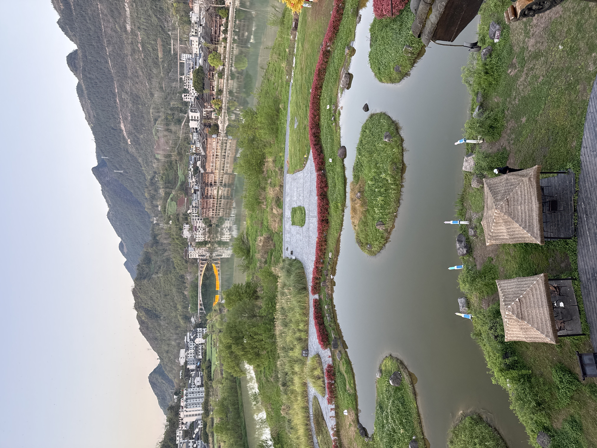 This aerial view shows a park-like setting in the city of Zhuoshui, China. A mountain range is visible in the distance, adjacent to a town reflected in a body of water. The park area features walking paths, landscaped areas with red flowers, and small islands in a pond. Two thatched-roof structures are present, with seating. Blue flotation devices are docked by the pond's edge. A golden bridge spans the water near the town. The sky is visible in the upper left corner.