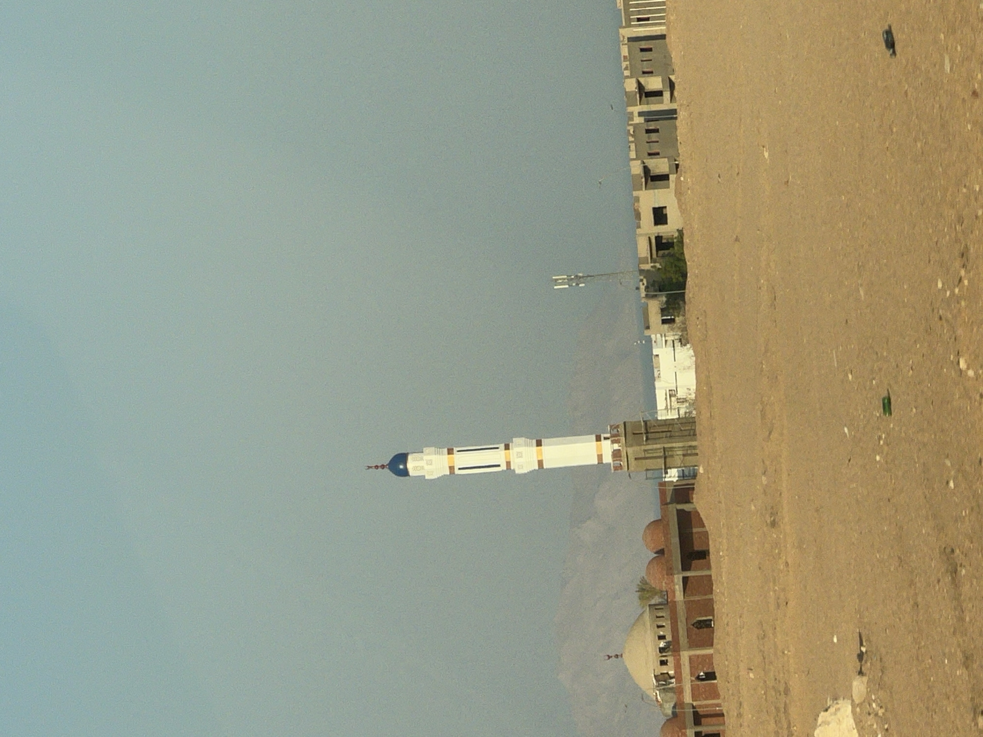 The image depicts a scene in Sharm El Sheikh, Egypt. In the foreground, a vast expanse of sand stretches across the frame, sparsely populated with small rocks and debris. A few green objects, possibly litter, are visible on the sand. In the mid-ground, a mosque complex is under construction. The main building features a large dome and several smaller domes, constructed with red brick. Adjacent to this, a tall, white minaret with blue and yellow accents stands prominently. Further in the background, a row of unfinished buildings with square openings for windows and doors lines the horizon. A metal tower with antenna-like structures is also visible. In the far background, hazy mountains create a backdrop against a clear, light blue sky. The overall environment suggests a desert landscape with ongoing development. There are no people visible in the image.