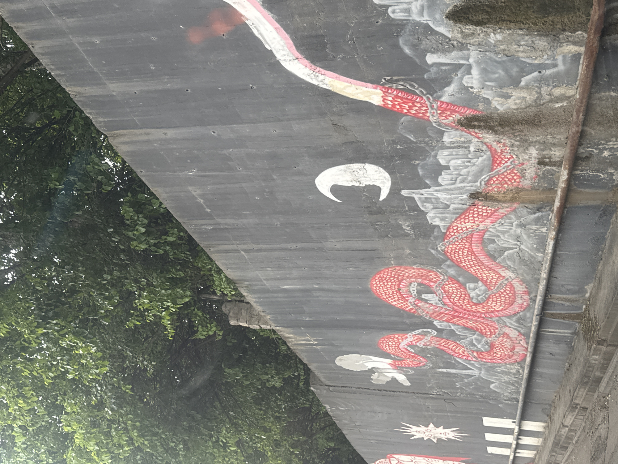 The image shows a low-angle view of a concrete overpass or bridge support structure adorned with graffiti-style artwork. The artwork depicts a large, stylized red and white snake winding across the surface. The snake appears to be shackled with chains in several places along its body. Other smaller white graffiti elements are present, including what appears to be a crescent moon shape. The concrete is dark gray. To the left, a dense mass of green foliage and tree branches fills a portion of the frame, suggesting a natural environment near the structure. A rusty metal pipe runs vertically along the right side of the overpass. Based on the reference information, this artwork is located in Tbilisi, Georgia.