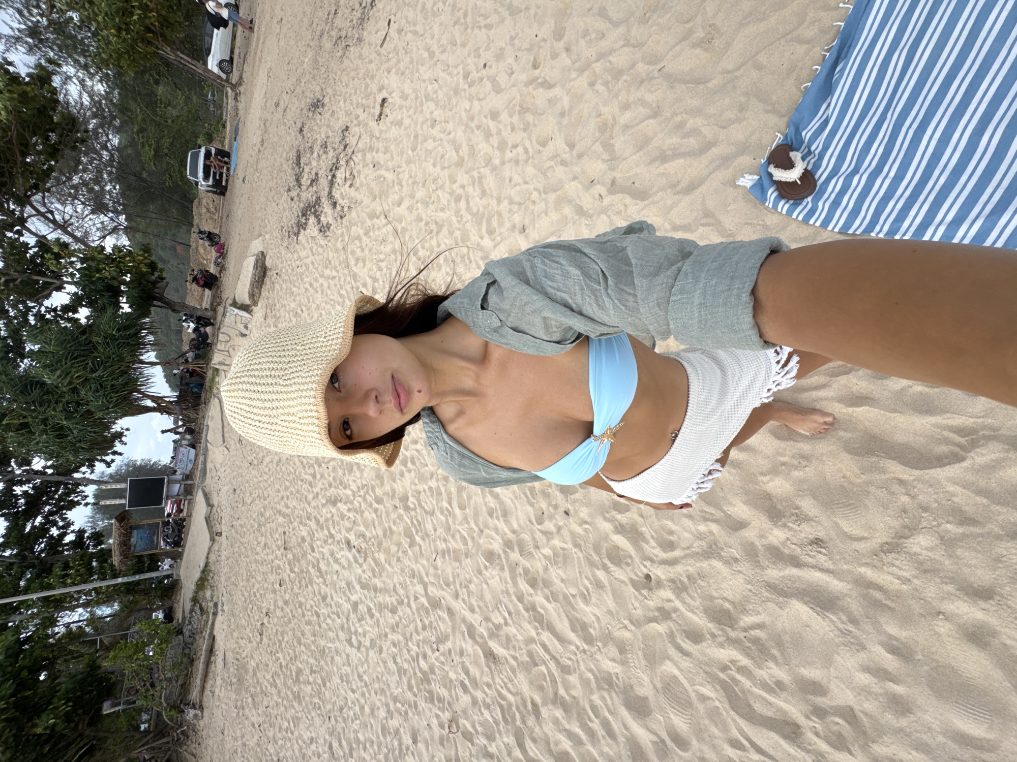 A woman wearing a straw hat, light blue bikini, white cover-up shorts, and an open light green shirt is the focus of this eye-level selfie. She is positioned on a sandy beach, likely in Rawai, Thailand, given the presence of tropical vegetation in the background and the coastal setting. The beach is occupied by trees and vegetation on one side, with a few parked vehicles, and people visible in the distance. In the right upper corner, there is a striped blue and white towel with a pair of sandals lying on it.