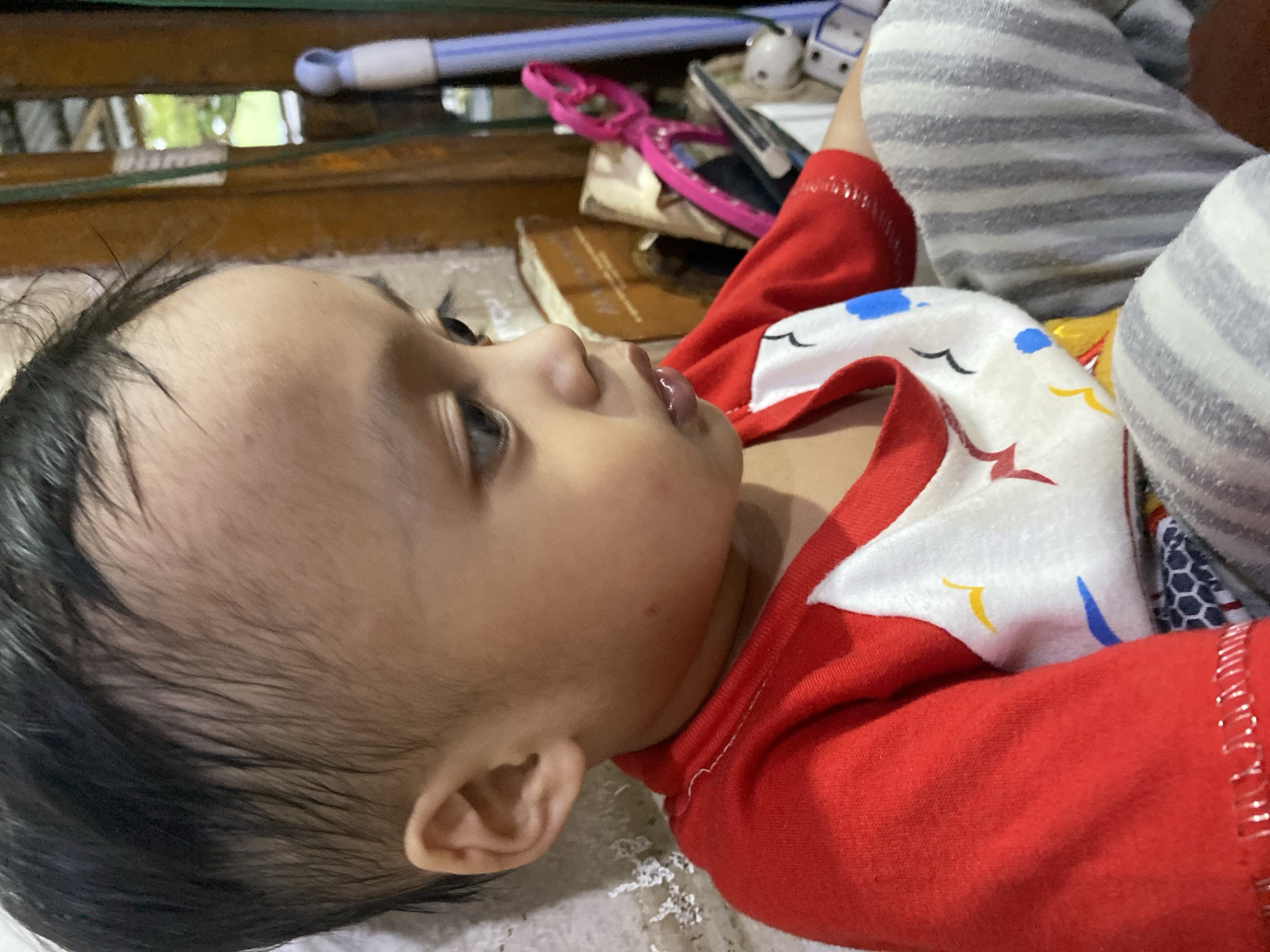 A young child is shown from the chest up, angled to the right. The child has dark hair and brown eyes. They are wearing a red shirt with a white bib that has colorful bird figures on it, and gray striped pants. The background appears to be indoors, with a wooden wall and some objects that suggest a storage or utility area. The lighting is dim. This image was taken in Ta', Indonesia.
