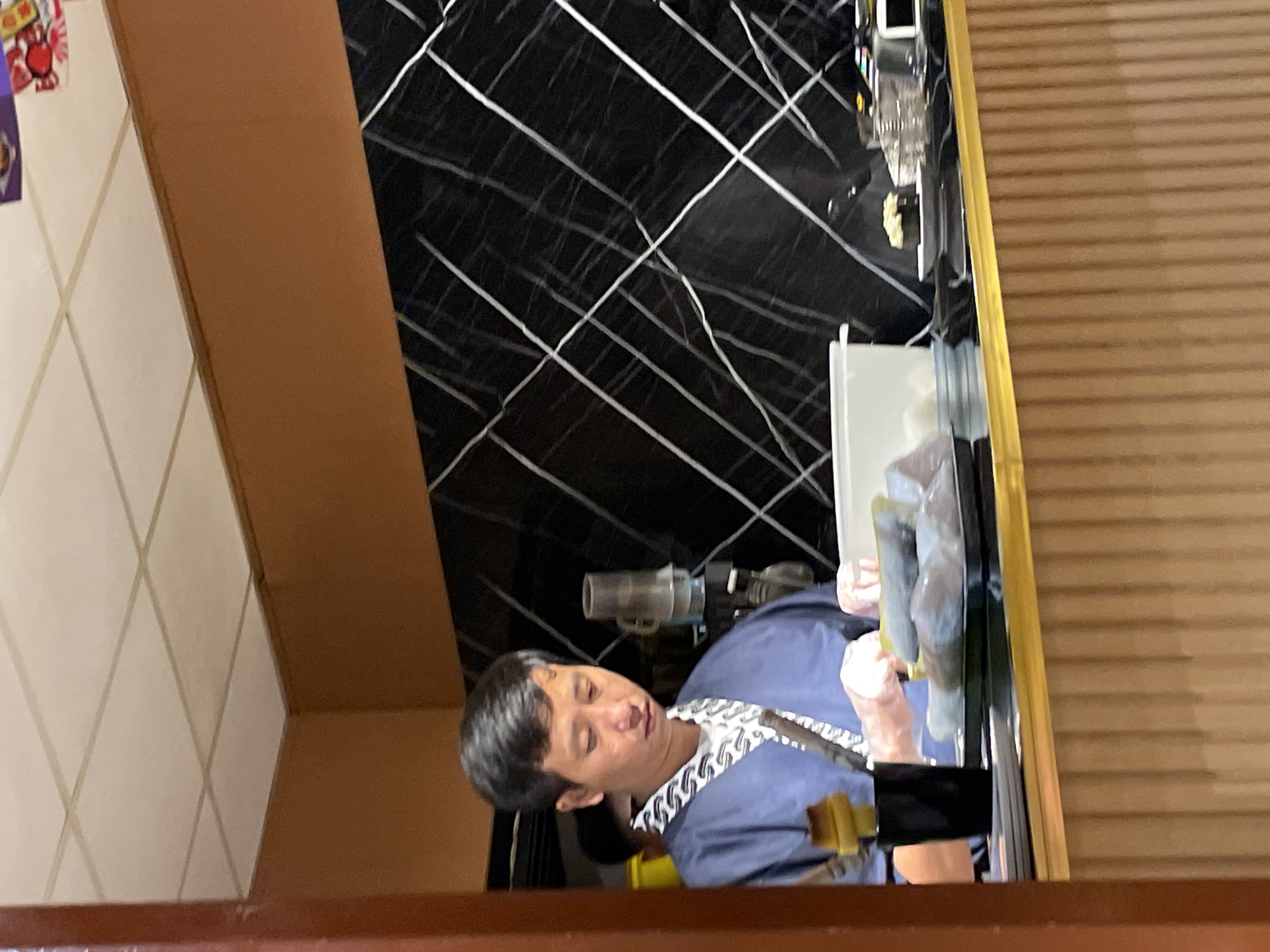 In Lâm Viên - Đà Lạt, Vietnam, a man wearing a dark blue traditional garment with a white patterned collar is preparing food behind a counter. He has short, dark hair and appears to be wearing pink gloves. In front of him, on a black tray, are several food items, possibly sushi or rolls, wrapped in translucent material. To his left, a small black cup holds a layered, golden-brown food item. The background features a black marble-patterned wall with white diagonal lines. The counter has a light brown wooden facade and a gold trim. Above the counter, a tiled ceiling is visible, and a portion of a brown wooden structure is on the left edge of the frame.