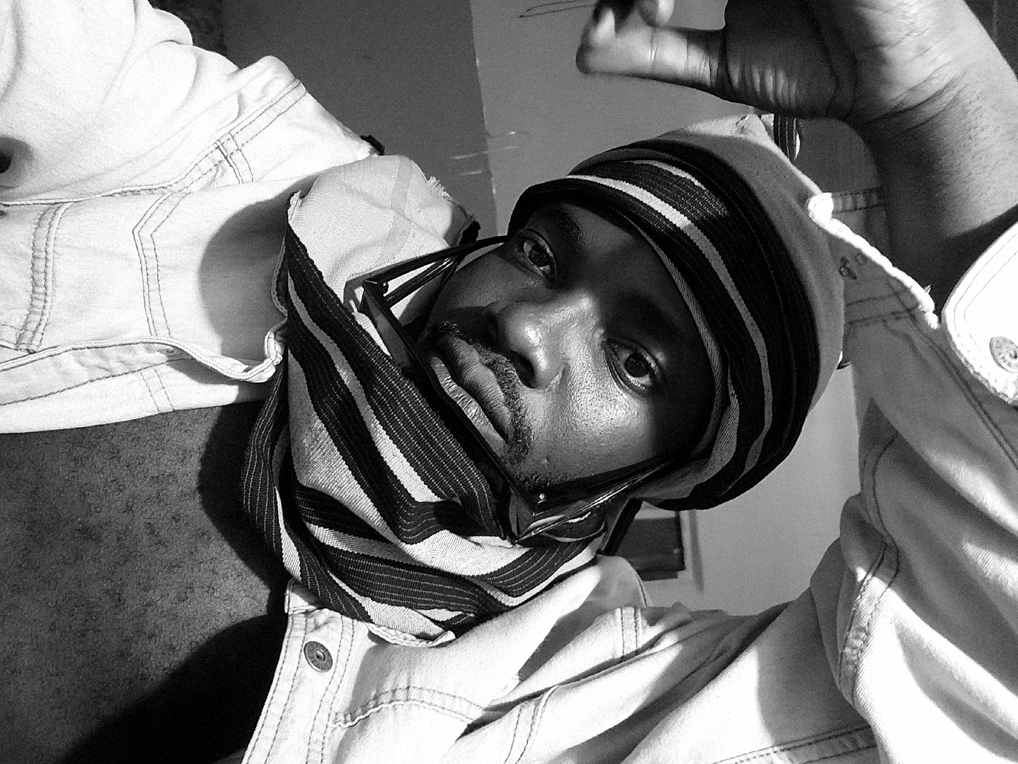 This high-contrast, black and white close-up portrait features a young Black man looking directly into the camera with an intense, serious expression. The shot is angled, suggesting a selfie or a low-angle perspective. He wears a striped head covering, possibly a beanie or hat, combined with a matching striped scarf or cowl that wraps around his neck. A pair of dark-framed glasses are positioned low on his nose, with the frames visible beneath his eyes. He has a neatly trimmed mustache and goatee.
He is dressed in a light-colored denim jacket, identifiable by its visible stitching and a metallic button on the right cuff that bears the faint inscription "ORIGINAL." His left hand is raised towards the upper right of the frame, with fingers slightly splayed, casting a soft shadow. The background is dark and largely out of focus, providing minimal contextual details about the scene. The image has a raw and direct feel, emphasizing the subject's gaze. The setting is Jos, Nigeria.