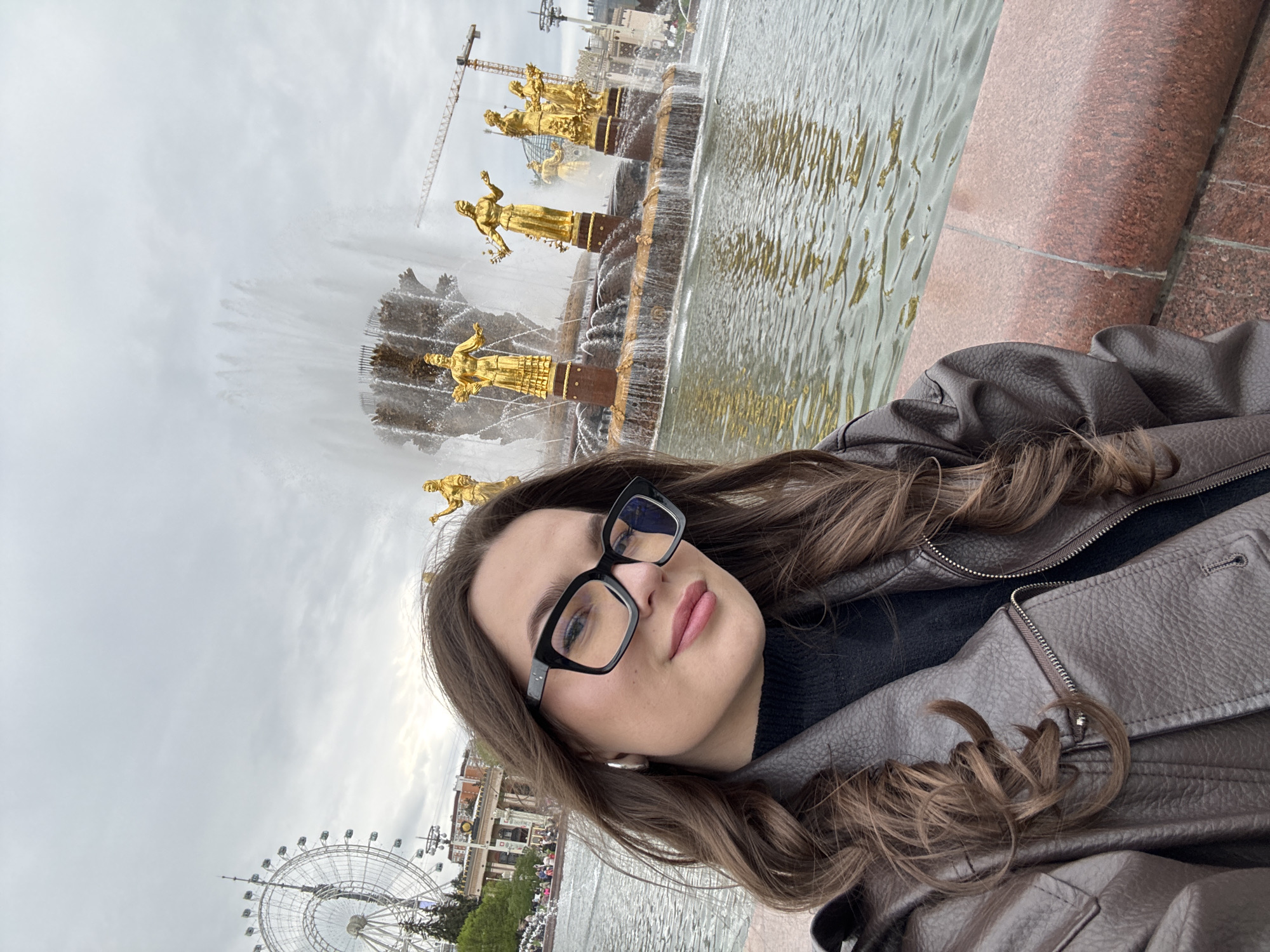 A woman is posing for a selfie. She is wearing a brown jacket and glasses. Behind her is the Friendship of Nations Fountain, a major landmark in Moscow, Russia. A Ferris wheel and a construction crane are also visible in the background. The weather appears cloudy.
