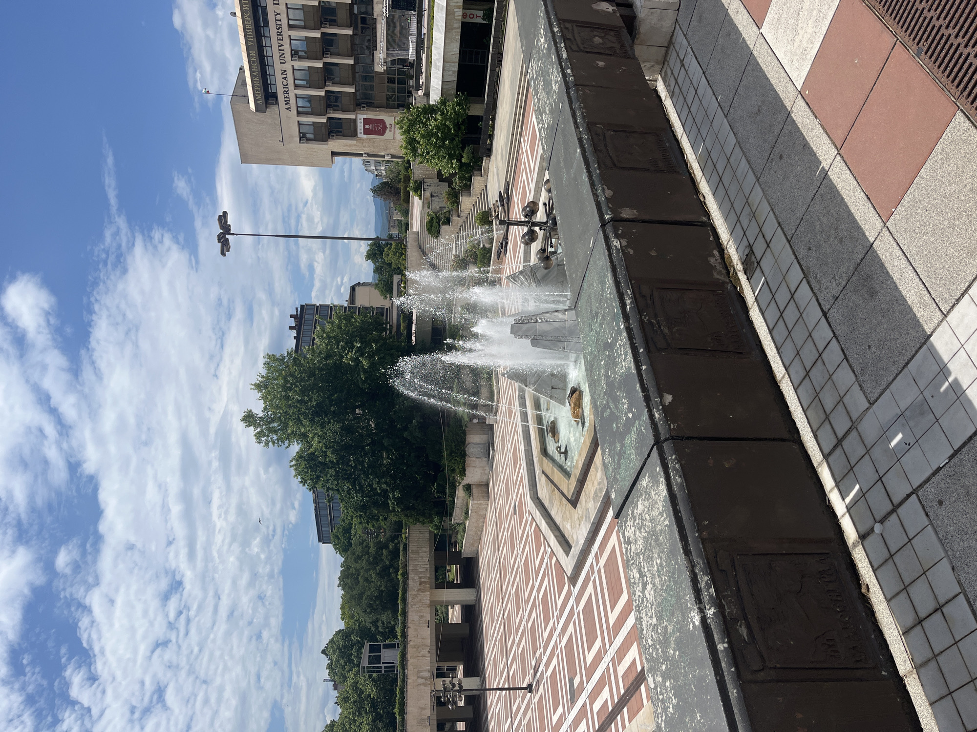 The image shows a cascading fountain in Blagoevgrad, Bulgaria. The fountain consists of multiple tiers and is located on a paved plaza. The pavers consist of red, brown and white color. In the background, there is a building with "AMERICAN UNIVERSITY IN" partially visible on the side. A light pole is present in the background. The sky is blue with white clouds. The fountain is actively spraying water, indicating it is in operation. A dark brown raised platform in the foreground adds depth to the scene.