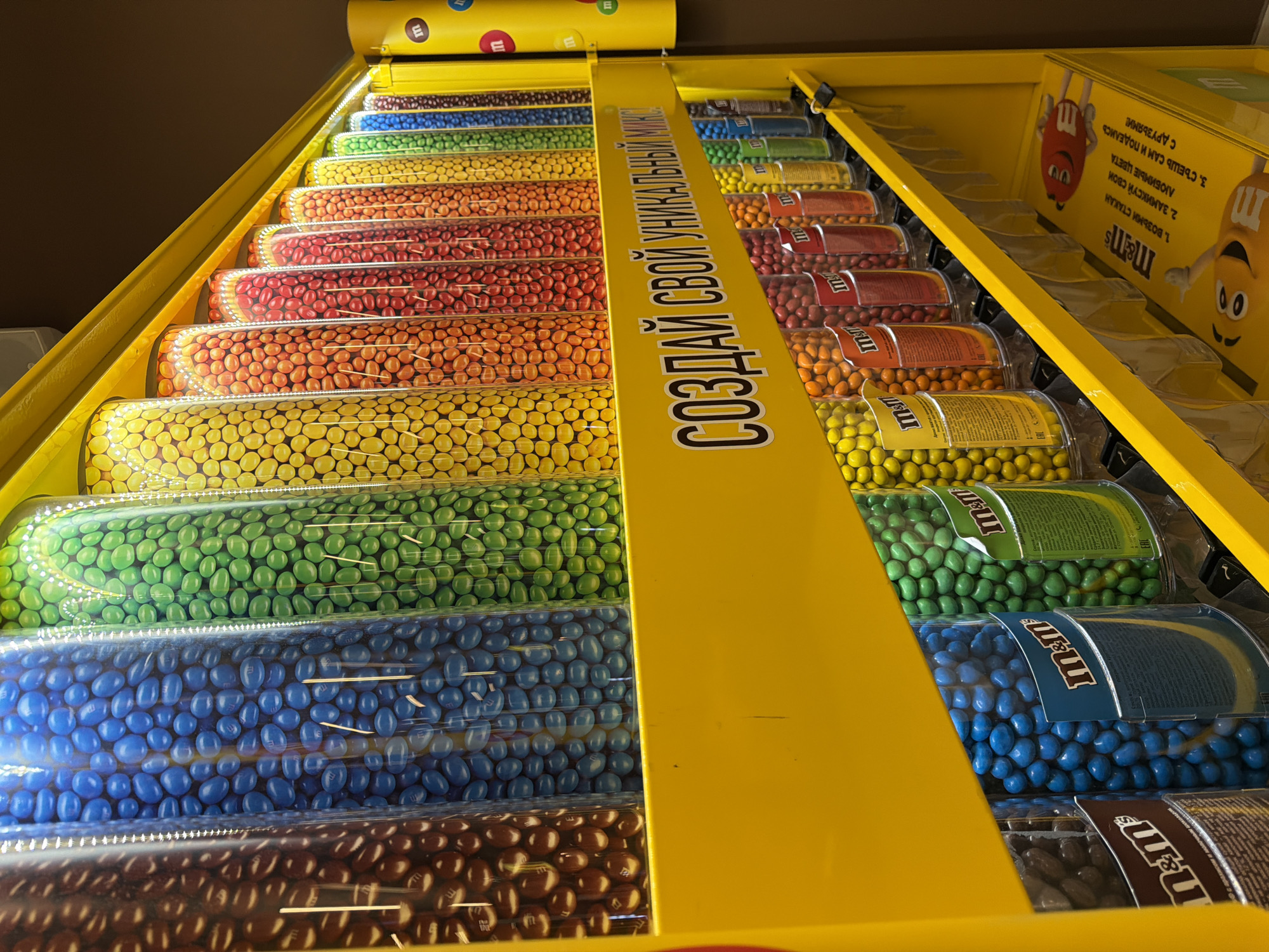 Here is a detailed description of the image:
The image shows a candy dispenser with a variety of M&M's separated by color, arranged in rows. The M&M's are neatly stacked and covered in clear packaging. The dispenser is yellow and has text in Russian that says "СОЗДАЙ СВОЙ УНИКАЛЬНЫЙ МИКС" which translates to "CREATE YOUR UNIQUE MIX". The dispenser appears to have instructions on how to use it, printed on the side with a cartoon M&M character. The colors of M&M's are arranged in a rainbow pattern. The exact location within the city of Unknown, Unknown cannot be determined from the image.