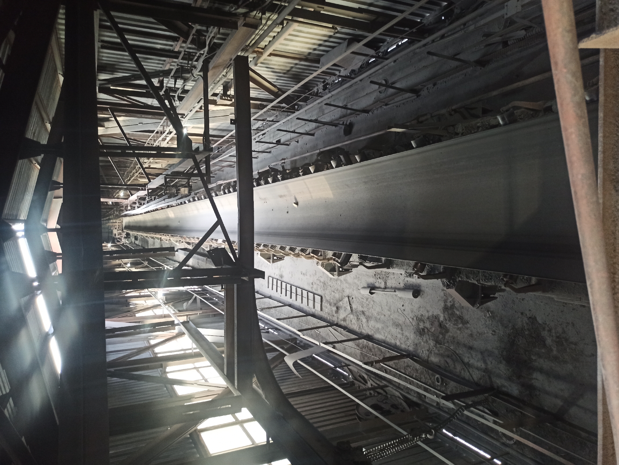 The image shows an interior view of an industrial facility, likely a conveyor belt system within a mine or processing plant in Kryvyi Rih, Ukraine. The scene is dominated by large metal structures, including beams, supports, and the extensive conveyor belt stretching into the distance. The lighting suggests it is daytime, with natural light filtering through corrugated metal roofing and windows, creating bright streaks and shadows. The overall atmosphere is utilitarian and somewhat aged, with dust and debris visible on surfaces and the floor. The conveyor belt itself is wide and appears to be made of a dark, durable material, with smaller mechanical components visible above and around it. There are no people visible in the image, and the focus is entirely on the industrial machinery and the structure of the building. The vast scale of the conveyor system is emphasized by the perspective, which draws the eye down its length towards a dimly lit, distant end. The presence of various pipes, wiring, and smaller machinery suggests an active, or recently active, industrial operation. The rough texture of the concrete floor and the worn appearance of the metalwork contribute to a sense of ruggedness and heavy use.