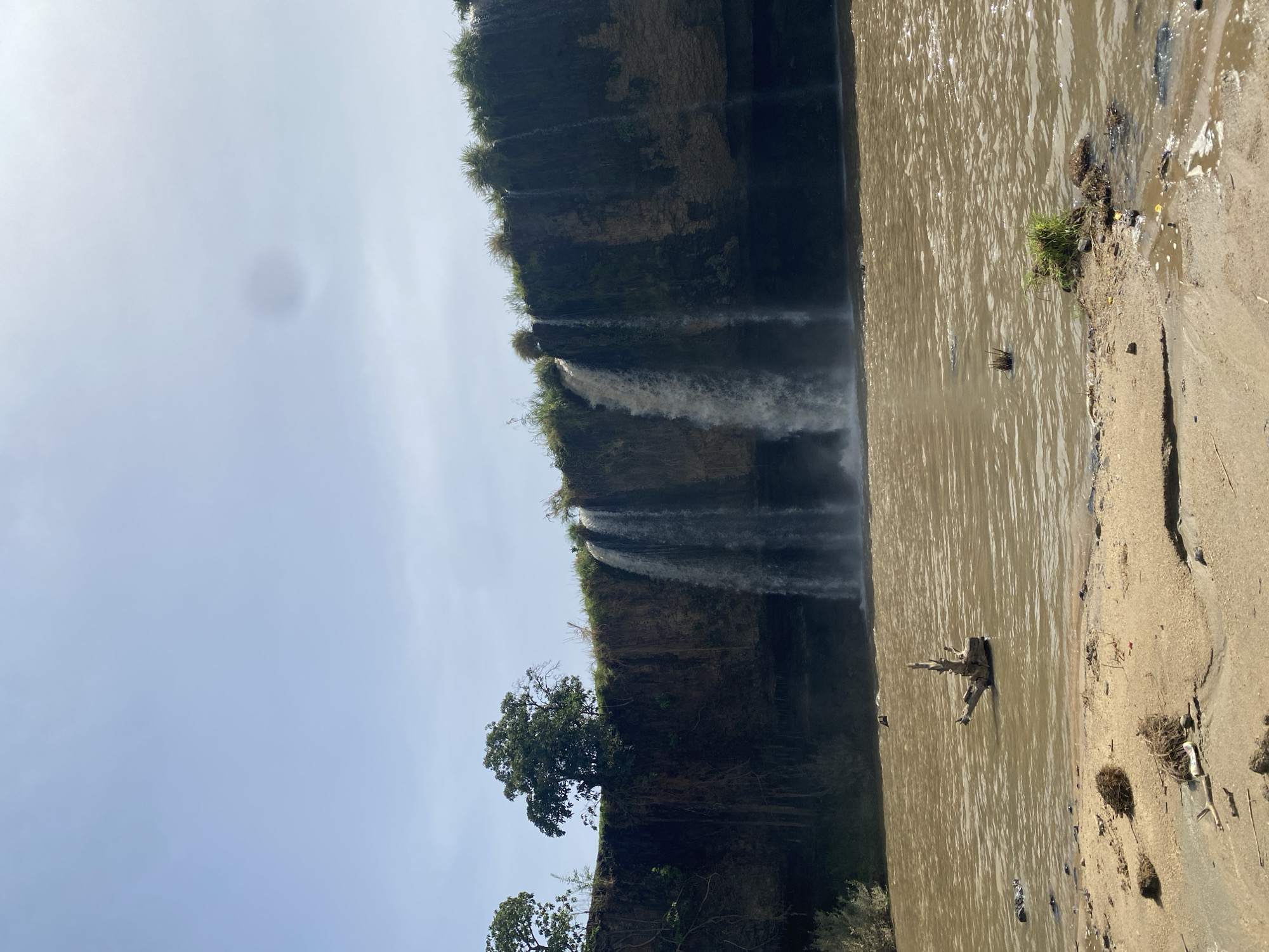This image depicts a natural landscape featuring a series of waterfalls flowing into a broad body of water, likely a river or lake. The scene is set in what appears to be a remote, vegetated area.
In the foreground on the right, a sandy or muddy bank slopes into the water. This bank is sparsely covered with small clumps of grass and scattered natural debris, including twigs and dried leaves, with some hints of possible litter. A prominent piece of driftwood or a partially submerged tree stump is visible in the water near this bank.
The central part of the image is dominated by a wide expanse of brownish, murky water, which reflects the bright sky above. Across this water, a tall, dark cliff face forms the background, densely covered in rich green vegetation, shrubs, and several large trees. Multiple streams of white water cascade down this cliff, creating several distinct waterfalls that plunge into the water below. One large tree stands out against the cliff face, towards the left of the waterfalls.
The sky, visible in the upper portion of the image, is bright and somewhat hazy, indicating daytime conditions, possibly under a high sun. A dark, circular smudge, likely a lens artifact, is noticeable in the sky.
There are no people or visible text in the image. The setting suggests a rugged, natural environment, consistent with the location context of Maigizo, Nigeria.