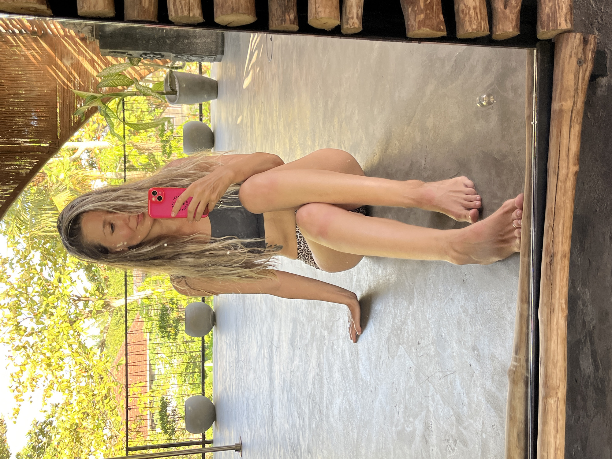 A woman with long blonde hair is taking a selfie in a mirror. She is wearing a dark grey sports bra and leopard print bikini bottoms. She is sitting on a concrete floor, with her legs bent and crossed. In the background, there is a lush green outdoor area with trees and a tiled roof structure. The environment appears to be a tropical setting, potentially in Weligama, Sri Lanka, given the architectural style and vegetation. The mirror is framed by rough-hewn wooden logs.