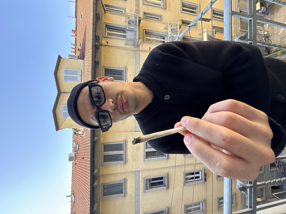 A male individual with dark hair, wearing a black beanie and black rectangular sunglasses, is depicted from the chest up on an urban balcony. He is dressed in a black long-sleeved buttoned cardigan. His left hand is extended towards the camera, holding a lit, partially consumed hand-rolled cigarette (joint) with a visible ash tip. The subject maintains direct eye contact with the camera, displaying a neutral expression.
The background features multi-story residential buildings consistent with architecture found in Milan, Italy. These structures have pale yellow stucco facades, numerous rectangular windows (some with light frames), and distinct dark blue/grey louvered shutters, many of which are closed on upper levels. The roofs are covered with terracotta tiles, displaying various vents and chimneys. An air conditioning unit is visible on a balcony of a building in the mid-ground. Below the balcony from which the image is taken, a glimpse of a street or courtyard reveals parked bicycles. The scene is illuminated by natural daylight under a clear, cloudless blue sky, indicating fair weather. The photograph is taken from an elevated position, likely several stories above ground level.