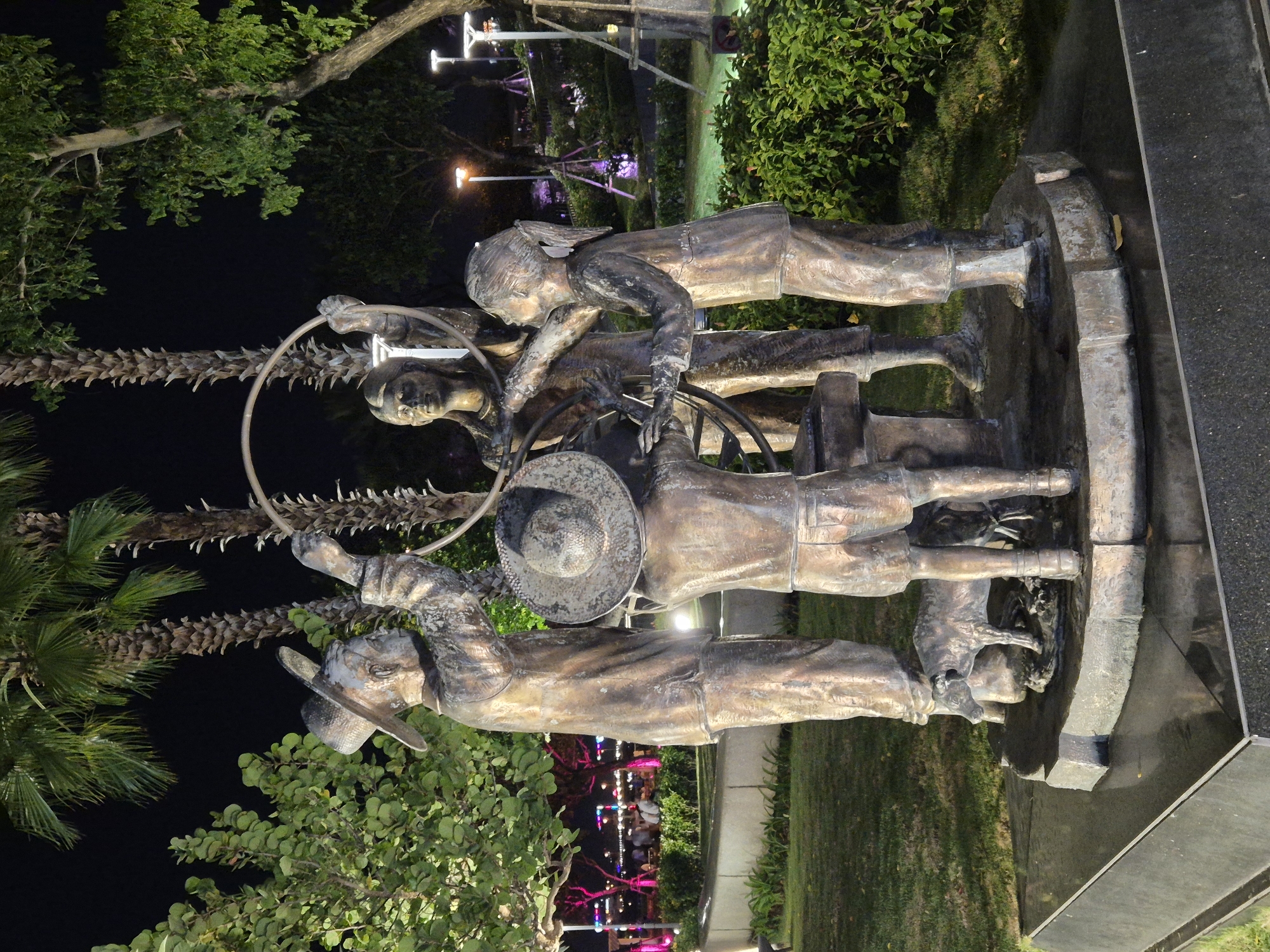 This is a nighttime photograph of a bronze sculpture depicting four children and a dog interacting with a large hoop. The sculpture is located in the city of Đà Nẵng, Vietnam, specifically at the East Sea Park. The children, dressed in what appear to be traditional Vietnamese clothing, are actively engaged with the hoop, suggesting a scene of play or communal activity. One child on the far left, wearing a hat, holds a part of the hoop. Another child in the center, facing away from the camera and also wearing a hat, seems to be assisting with the hoop. Two other children are also involved, one on the right reaching for the hoop and another to their left. A small dog stands at the base of the sculpture, looking towards the children. The sculpture is set within a well-lit park environment, with lush green bushes and trees surrounding it. In the background, palm trees are visible, and further in the distance, there are illuminated buildings and streetlights, indicating an urban setting. The lighting in the park highlights the sculpture and the surrounding greenery, creating a dramatic effect in the darkness.