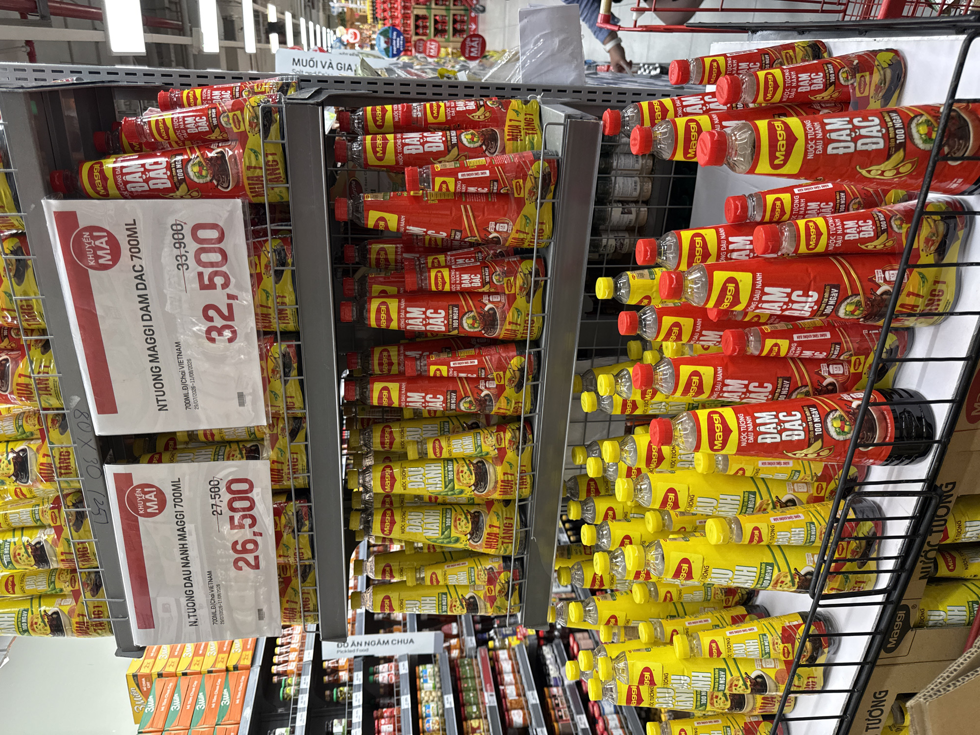 The image shows a well-lit supermarket aisle in Nha Trang, Vietnam, with a focus on a display of Maggi brand sauces. The shelves are stocked with various Maggi products, predominantly soy sauce and a dark, thick sauce. Prominent signage indicates a sale, with prices clearly marked. One sign advertises "N. TUONG DAU NANH MAGGI 700ML" with a discounted price of 26,500 VND, down from 27,500 VND. Another sign promotes "NTUONG MAGGI DAM DAC 700ML" with a price reduction from 33,900 VND to 32,500 VND. Bottles of sauce are arranged in rows on metal shelves, with several bottles in the foreground featuring prominent red and yellow labeling. In the background, other grocery items are visible on shelves, and a red shopping cart is partially seen on the right side of the image. A person's arm, wearing a blue sleeve, is visible near the shopping cart. The environment suggests a typical retail setting with clear organization of products.