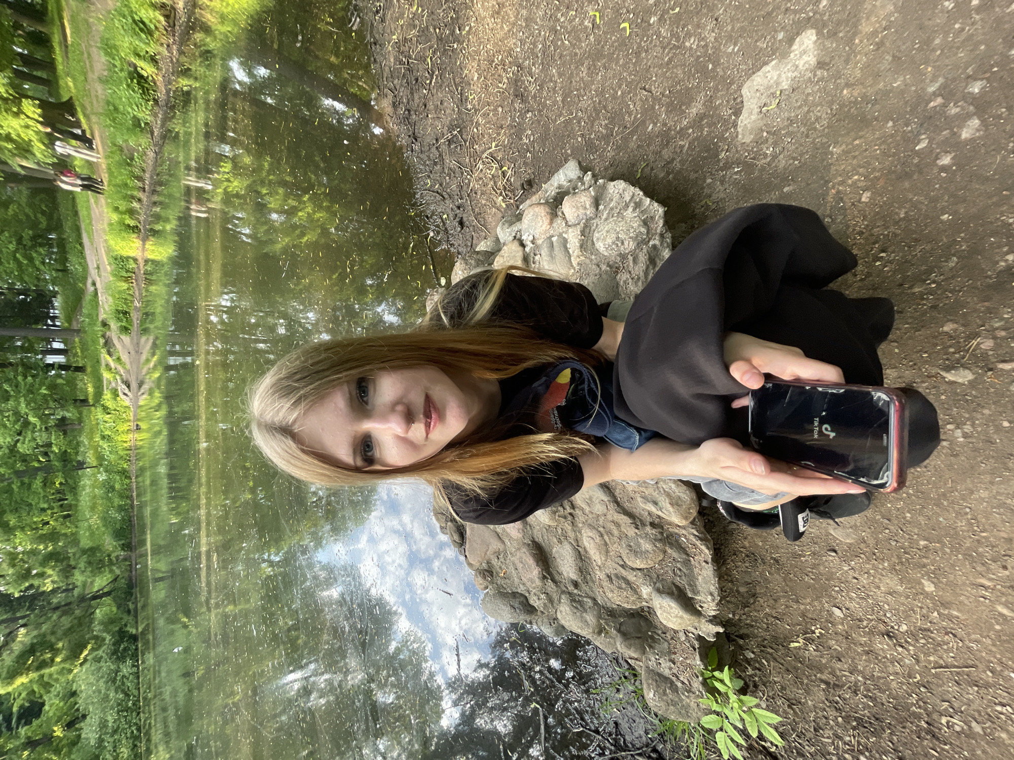 A young woman with long, blonde to light brown hair and blue eyes is pictured outdoors, likely in a park in Saint Petersburg, Russia. She has a nose piercing and is wearing a black top, a denim jacket, and dark bottoms. She appears to be sitting or crouching on a rocky bank beside a body of water, possibly a pond or lake. The ground consists of damp earth, mud, and scattered debris near the water's edge, transitioning to drier dirt further away.
The woman looks directly at the camera with a calm, neutral expression. She holds a black smartphone in her hands; its screen is visibly cracked, and the "TikTok" logo is clearly displayed on it.
The water's surface reflects the surrounding dense green trees, overhead sky with clouds, and distant light. Across the water, lush green foliage and trees line what appears to be a walking path, with indistinct shapes that might be benches or other park visitors. The lighting suggests it is daytime, possibly during the late morning or afternoon, with dappled sunlight filtering through the leaves. The atmosphere is peaceful, capturing a candid moment in a natural, urban park environment.
