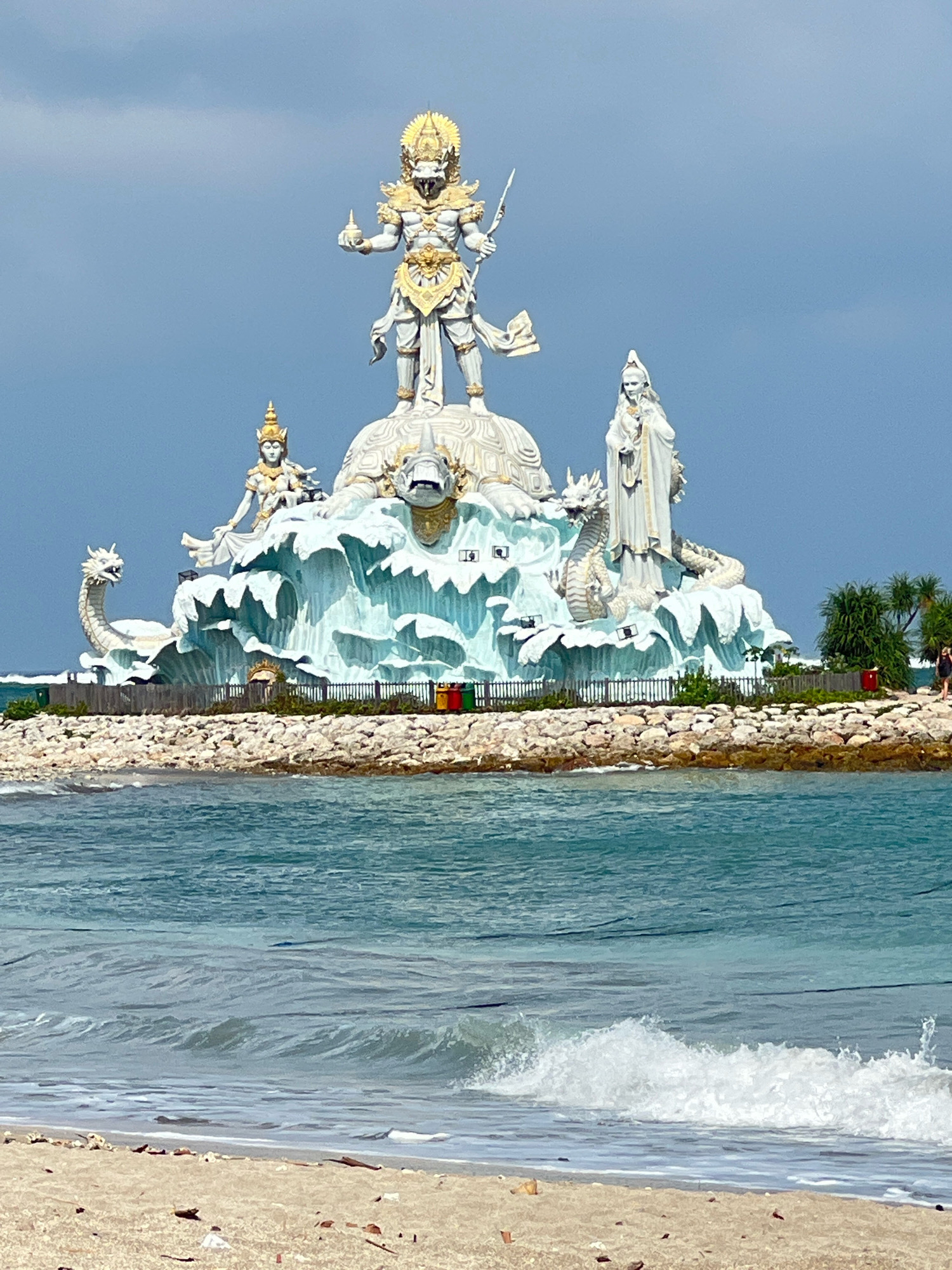 The image depicts the Garuda Wisnu Kencana Cultural Park in Kuta, Indonesia. The colossal statue of Vishnu riding Garuda dominates the center, rendered in white and gold. To the left, a seated figure with a headdress sits on a large, stylized wave formation, accompanied by a white dragon. To the right, a standing figure in flowing robes holds a staff, also positioned on the wave structure, with another white dragon coiled nearby. The statues are set on a rocky outcrop, with a fence and some vegetation visible behind them. The sea in the foreground is a vibrant turquoise, with gentle waves breaking on a sandy beach. The sky is overcast with a bluish-gray hue. A few people are visible in the distance on the right side of the image, walking near the park's edge.