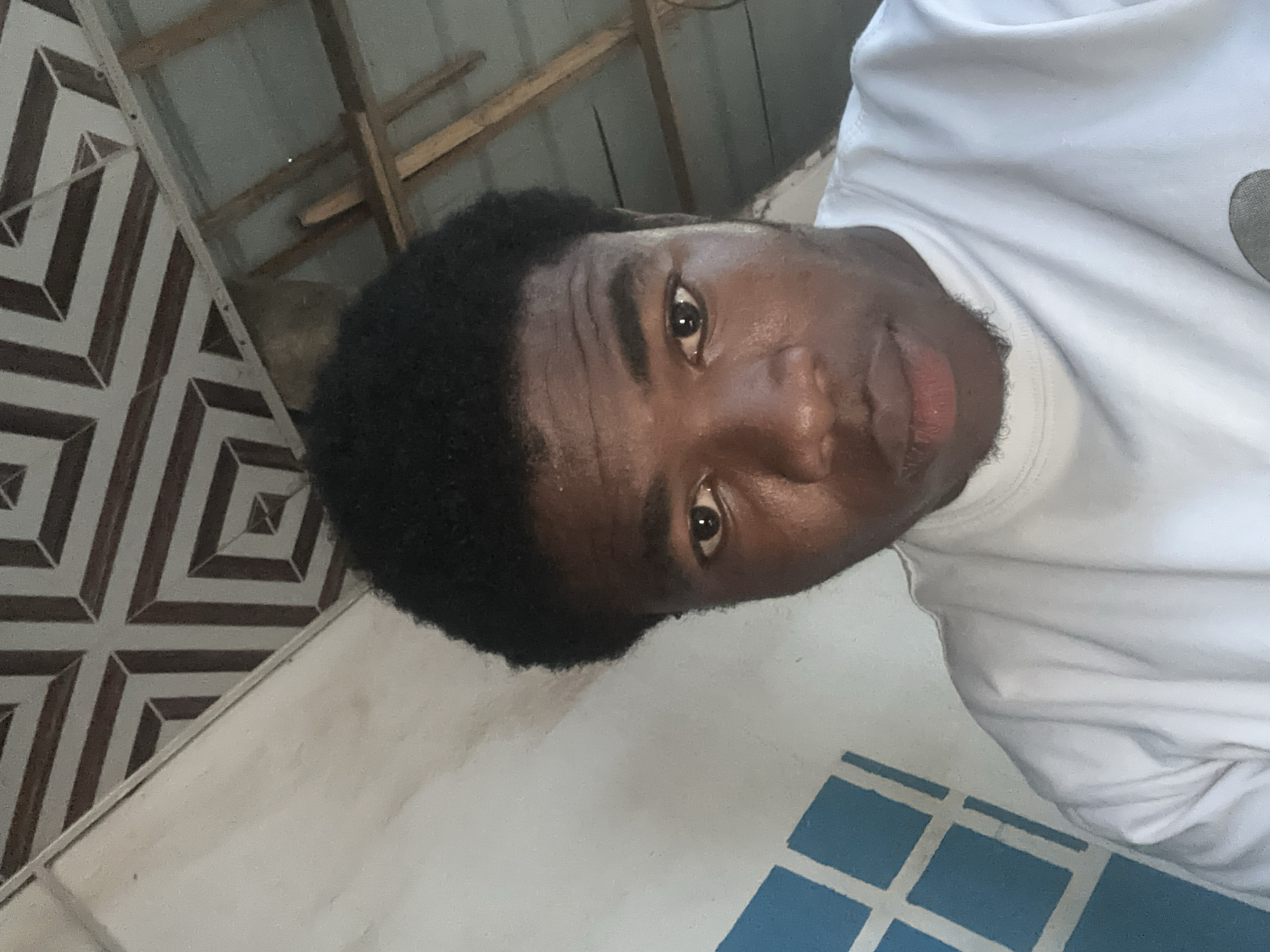 The image shows a person with dark skin and short, black hair wearing a white t-shirt. The person is looking directly at the camera. Behind the person, the background includes tiled walls with geometric patterns and a ceiling with wooden beams. A wall with blue square designs is visible. The location is Orie Uratta, Nigeria.