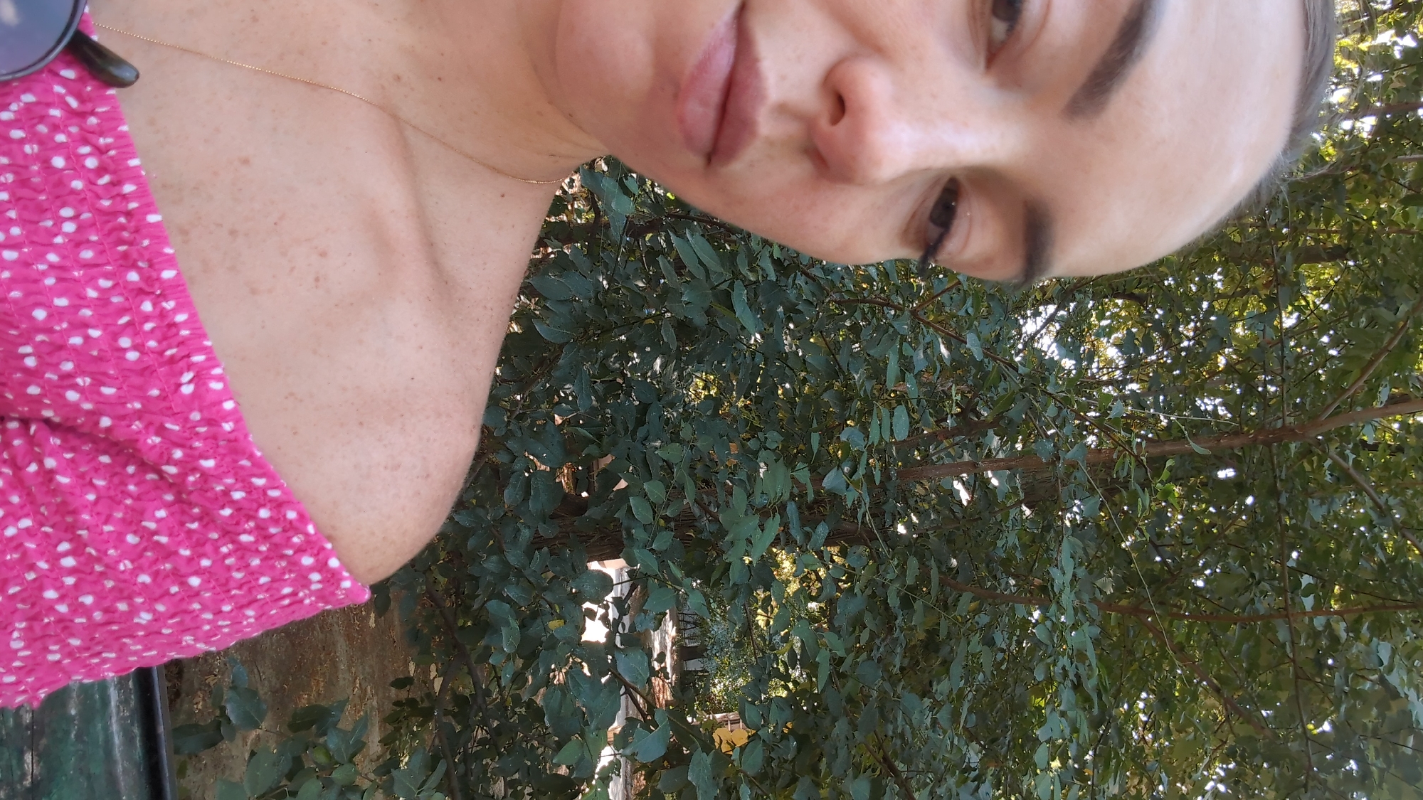 A woman is pictured from the chest up, her body turned to the left. She has fair skin with freckles and is wearing a pink off-the-shoulder top with white polka dots. A thin gold chain necklace is visible. The background consists of green foliage from trees. The image was taken outdoors, likely in a park or garden, with dappled sunlight filtering through the leaves. There is no identifiable location in Chisinau, Moldova.