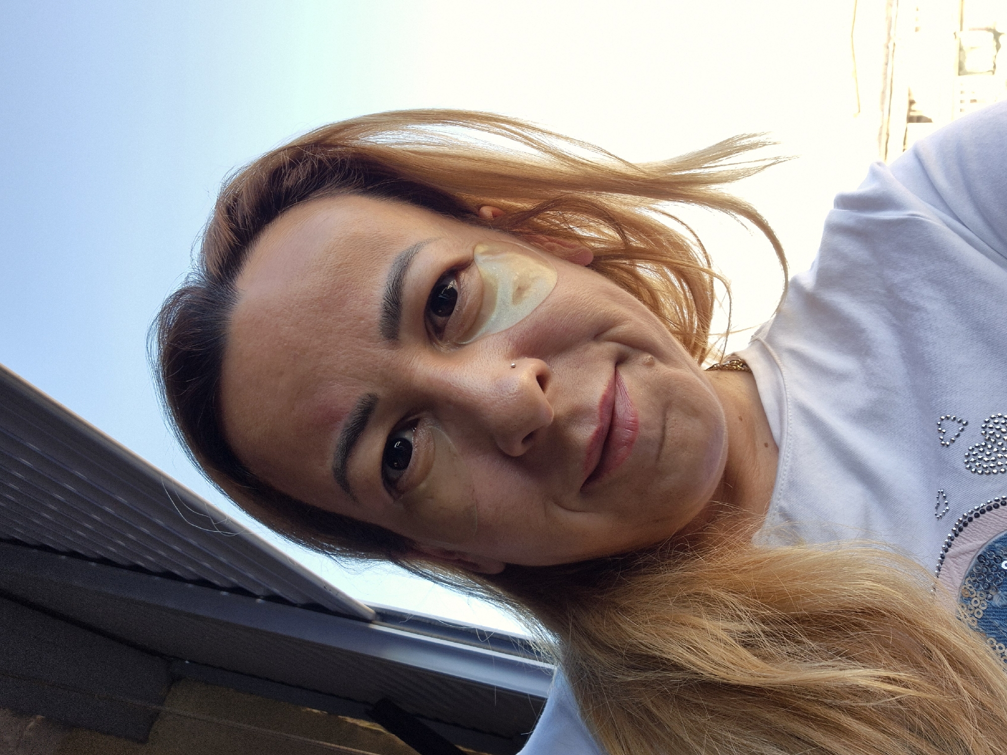A medium shot taken from below, captures a woman from the shoulders up as she looks directly at the camera. She has long, blonde hair that is slightly wavy and falls over her left shoulder. She is wearing a white t-shirt with a rounded neckline. There are two light-colored eye patches on her cheeks, below her eyes. Her eyebrows are dark and arched, and she has a small nose ring. The background shows a bright blue sky and what appears to be an open window with blinds. The lighting suggests it is daytime, and the overall scene implies an indoor or outdoor setting near a window.