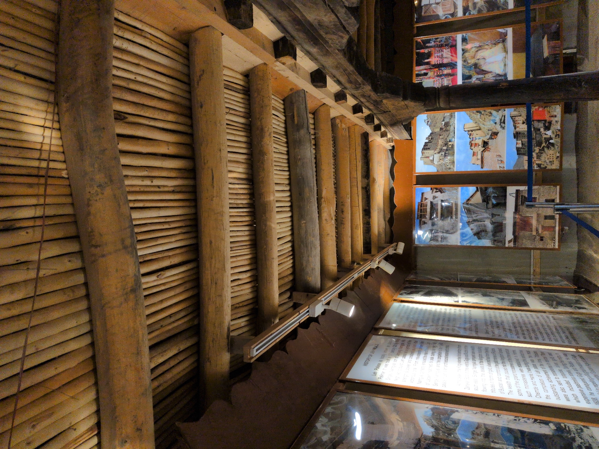 The image shows the interior ceiling of a building, likely in Leh, India, constructed with rough-hewn wooden beams and a layer of smaller wooden poles beneath. The ceiling is angled, creating a sense of depth. Below the ceiling, on the left side of the image, are several large framed displays with text and images. One display, labeled "HISTORY OF LEH PALACE," contains text detailing the history of the Leh Palace. To the right of these displays are further framed pictures, some showing architectural scenes and others depicting what appear to be historical or cultural subjects. The lighting in the room comes from ceiling-mounted track lights, one of which is clearly visible and illuminated. In the foreground, on the right, a thick, dark wooden support post is prominent. The overall impression is of an exhibition space within a historic structure, with informational panels and photographic displays. The architecture suggests a traditional building, possibly a museum or visitor center related to local history.