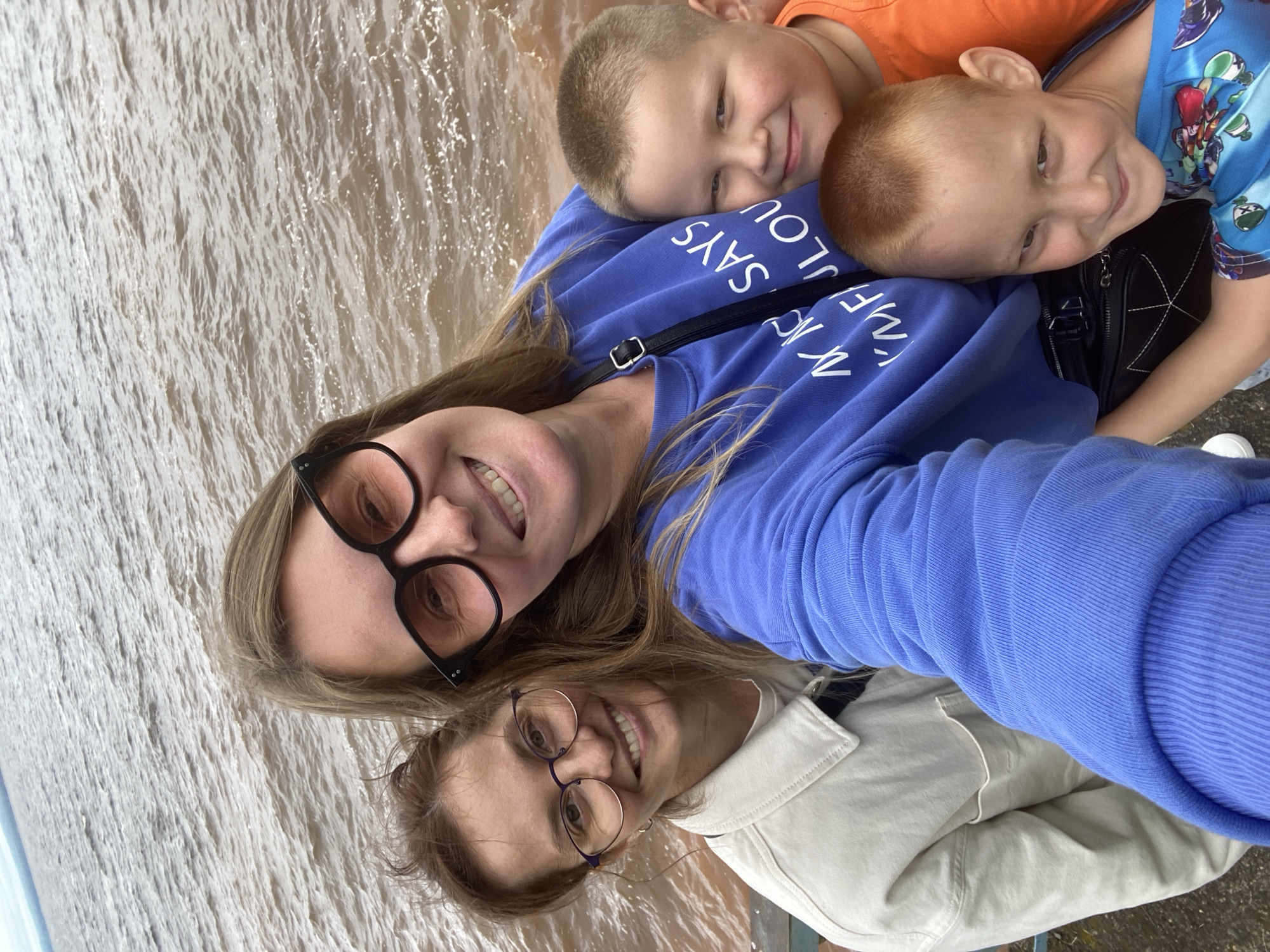 The image shows a group of four people taking a selfie outdoors, with a body of water in the background. Two women are in the foreground, smiling and looking at the camera. One woman is wearing glasses and a light-colored jacket, while the other is wearing large-rimmed glasses and a blue sweatshirt with white text. Two young boys are positioned in front of the women, also smiling. One boy is wearing an orange t-shirt, and the other is wearing a blue t-shirt with a cartoon print. The water appears to be choppy and a brownish color, suggesting it may be a river or a muddy body of water. The environment appears to be an open area, possibly a pier or a waterfront promenade. There is no visible indication that the location is in Noviy, Russia.