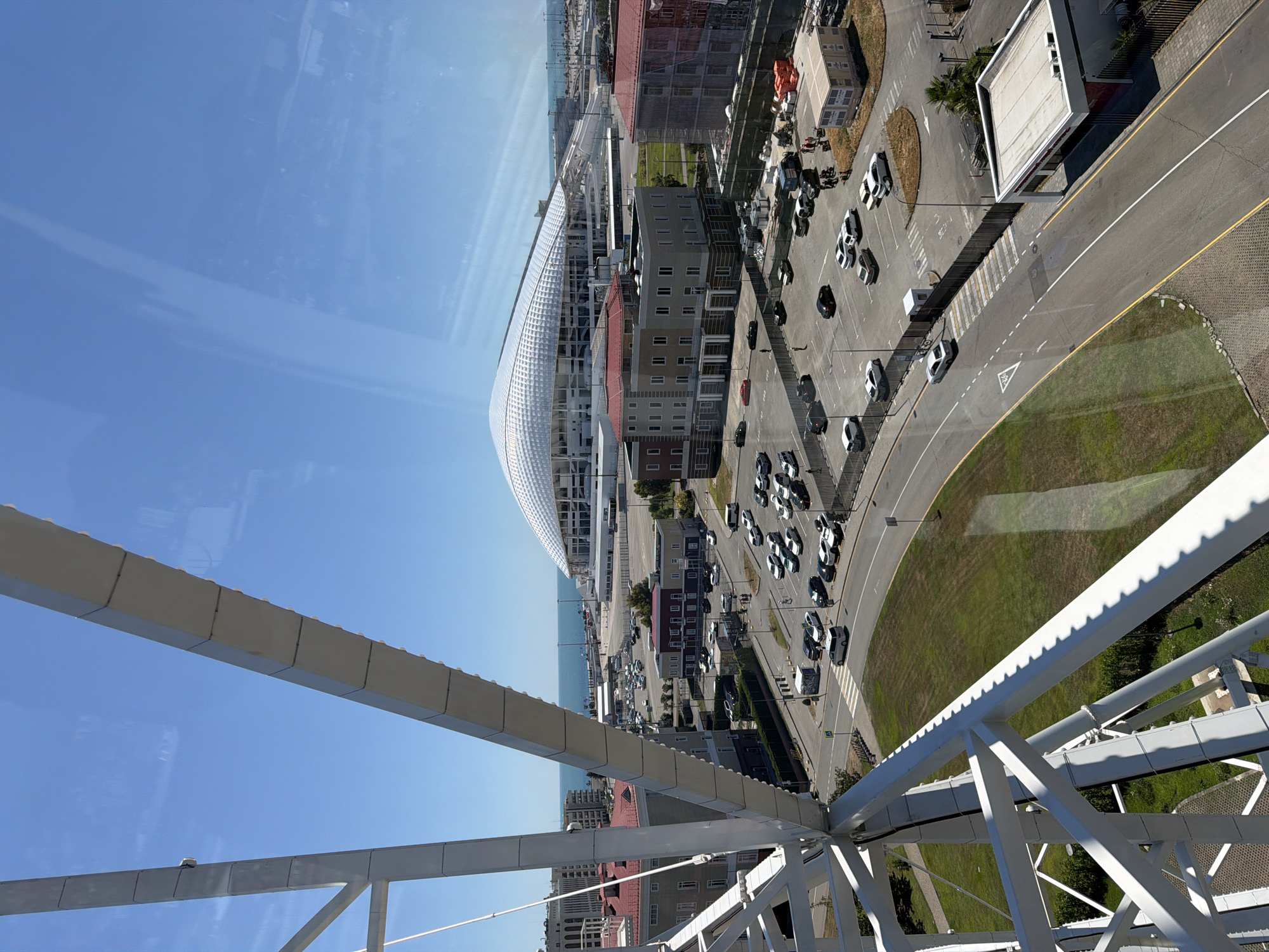The image shows an aerial view of a modern urban area with a distinctive white, curved building resembling a stadium or arena. The building is located near a body of water, likely the sea, suggested by a distant coastline and a clear blue sky. Several multi-story buildings, some with red roofs, are clustered around the main structure. A road with parked cars is visible, indicating a public or commercial area. The perspective suggests the photograph was taken from a high vantage point, possibly a Ferris wheel or a tall building, as evidenced by the white metal framework in the foreground, which is a structural element of the vantage point. The scene appears to be bathed in bright sunlight, suggesting a clear day. The presence of cars and buildings implies a developed urban environment, possibly a tourist or recreational area given the unique architecture and proximity to the sea. There is no visible text in the image. The overall impression is one of a vibrant, modern city by the coast on a sunny day.