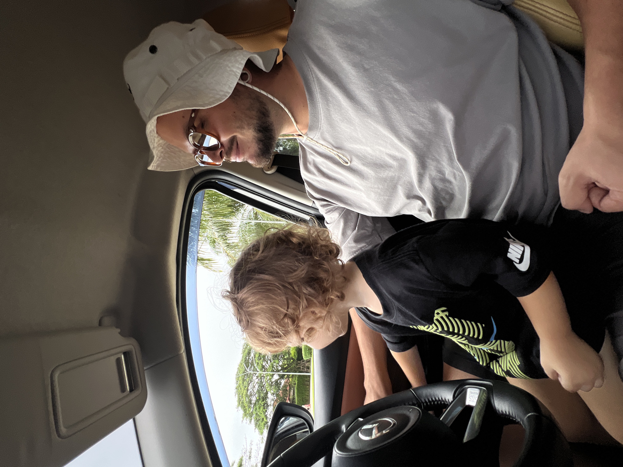 A father and son are seated in the driver's seat of a car. The father, wearing a white bucket hat and glasses, looks down at the steering wheel. The son, in a black Nike t-shirt, leans towards the steering wheel. The car is parked in an unknown location. Trees and the sky are visible through the car's window.