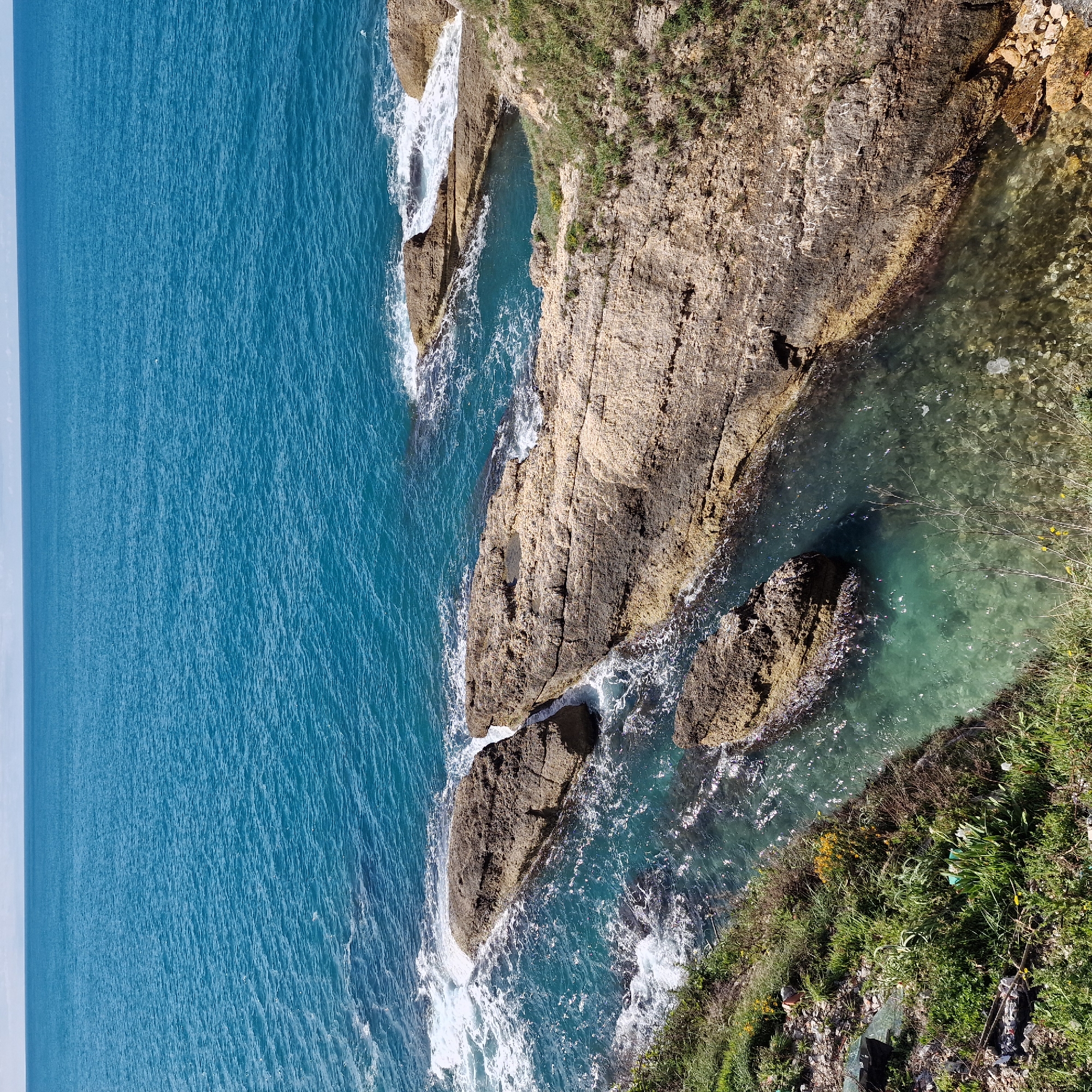 An aerial view of rocky shoreline in Ulcinj-Ulqin, Montenegro. A deep blue sea meets a rugged coastline of light brown rocks. White foam from crashing waves is visible around the rocks. Patches of green vegetation grow on the rocks near the edge of the coastline. Water is visible within a small inlet between the rocks. The sky is obscured by the aerial viewpoint.