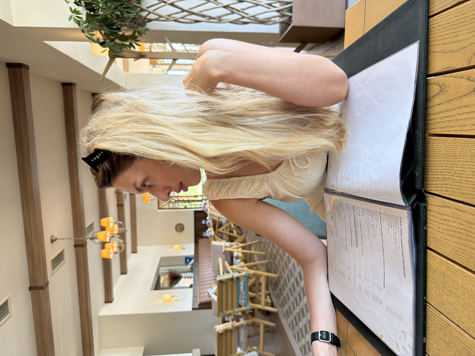 A woman with long blonde hair, wearing a cream-colored top and a black watch, is sitting at a wooden table in a restaurant. She is looking to her right, with her right arm resting on a menu. The restaurant appears to be in Bigor, Montenegro, with a bright and airy ambiance, featuring wooden beams on the ceiling, ornate chandeliers, and upholstered chairs. A tall plant in a pot is visible to the right of the woman, and other tables and seating areas are in the background. The menu in front of her is open, displaying various items and prices in what seems to be a local language. The lighting is bright, suggesting it is daytime.