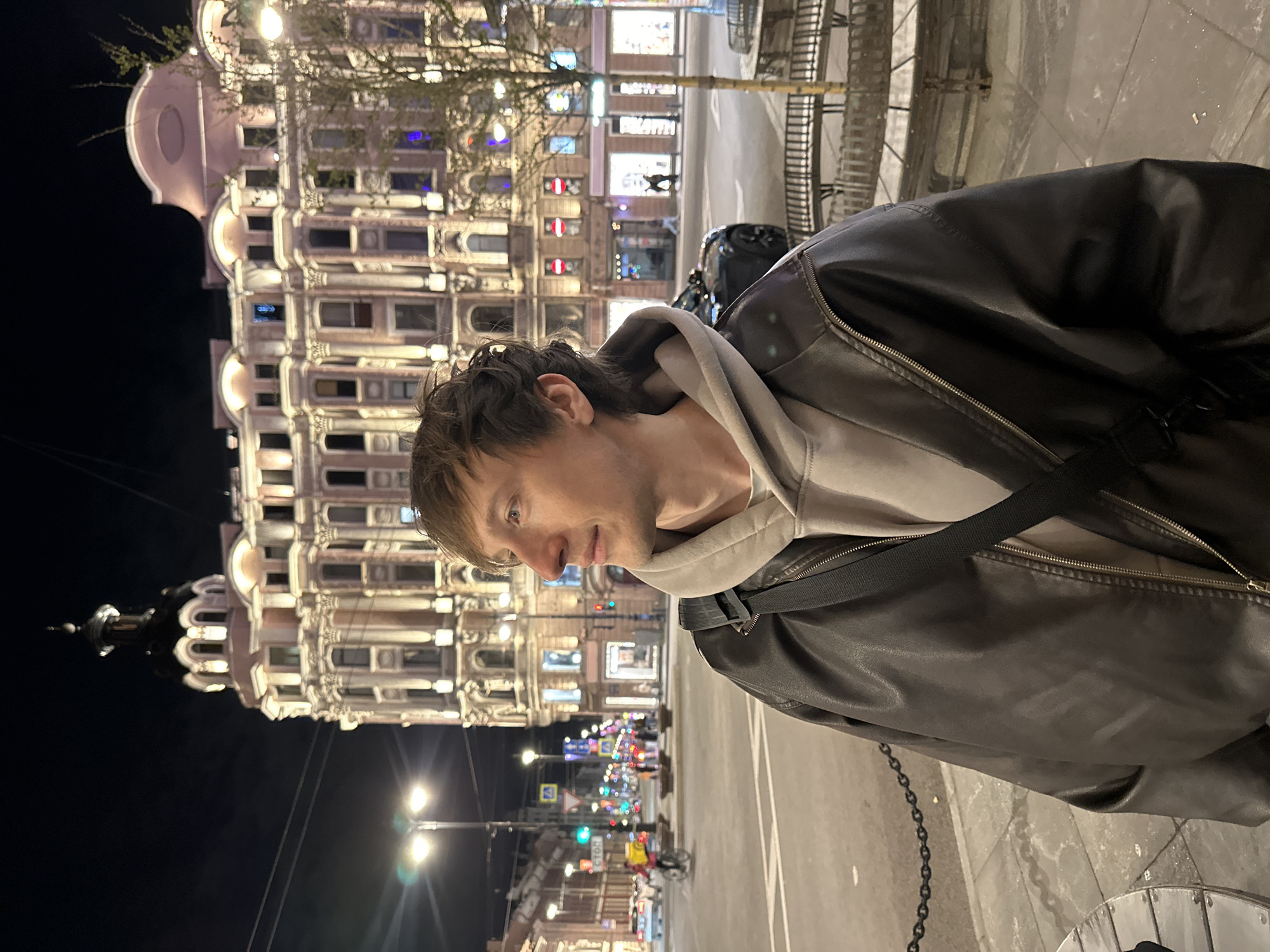 A vertical shot, rotated 90 degrees clockwise, captures a man standing on a city street at night. The man, with short brown hair and light eyes, is in the foreground, facing slightly to his left and looking downwards with a neutral, contemplative expression. He wears a dark brown or black leather jacket over a light grey hooded sweatshirt, with a dark strap of a bag visible across his chest.
Behind him, a large, ornate building dominates the midground, brightly illuminated against the dark night sky. The building exhibits classical architectural elements such as columns, intricate window frames, and decorative friezes, appearing grand and historic. Bare tree branches are visible in front of parts of the building.
The scene suggests an urban environment. Streetlights cast glows on the pavement and buildings. In the background, a street or road is visible with car lights, traffic signals (red and green lights are discernible), and the illuminated storefronts of other buildings. A bicycle and other indistinct figures can be seen in the distance on the street. A black chain runs along the bottom right of the frame, possibly part of a barrier.
The time of day is clearly night, with artificial lights providing the primary illumination. A visible street sign in the background features the Cyrillic word "СТОП" (STOP). Based on the architectural style and the provided context, the image is set in Saint Petersburg, Russia.