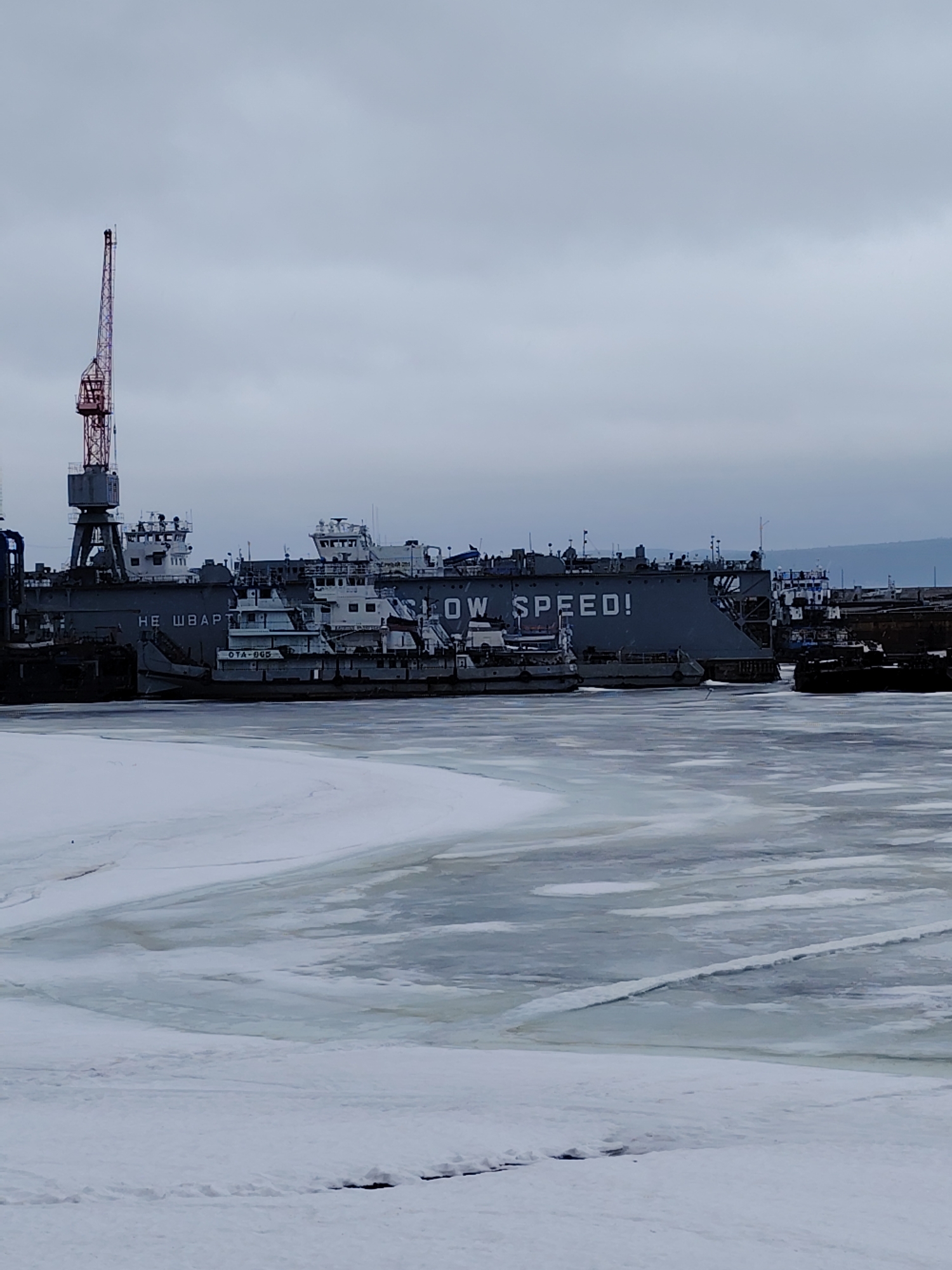 The image depicts a frozen harbor in Chaikovsky, Russia, on an overcast day. The foreground shows a mostly frozen body of water with areas of cracked ice and patches of snow cover. Several gray ships, some bearing Russian text, are docked along the harbor's edge. One ship prominently displays the phrase "LOW SPEED!" in large, white capital letters. A red and gray crane stands at the edge of the harbor. The sky is uniformly gray and overcast, contributing to the cold, industrial atmosphere of the scene.