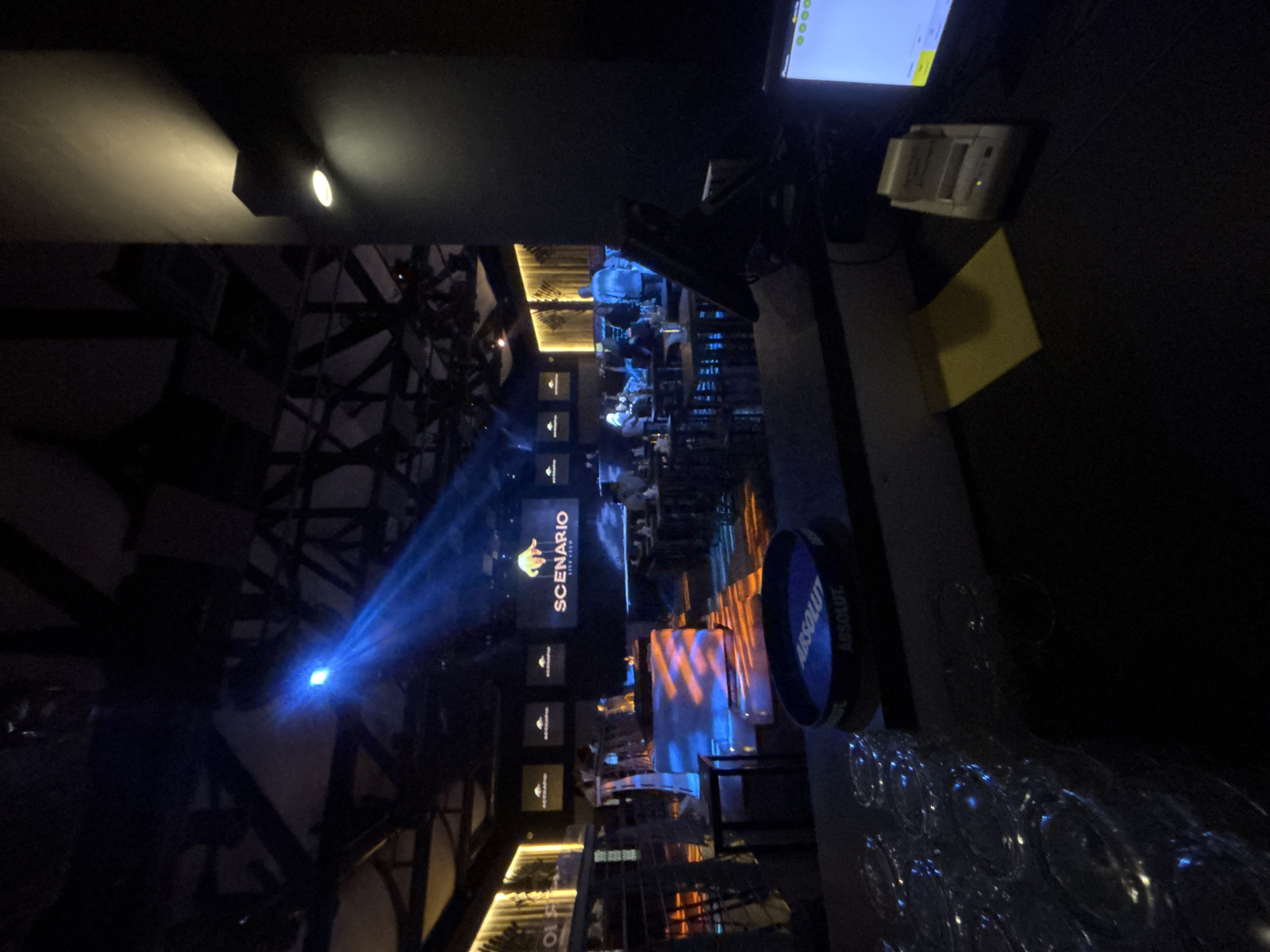 This image depicts the interior of a nightclub or bar named "SCENARIO LIVE CLUB" in Tsarevo, Bulgaria. The scene is dimly lit, with a prominent blue spotlight illuminating a section of the ceiling structure and casting a beam across the room. In the foreground, a bar area is visible, with a stack of glasses on the left and an "ABSOLUT" branded coaster on a dark surface. Behind the bar, tables and chairs are arranged for patrons, some of whom are seated and engaged in conversation. A large screen displays the club's name and logo. The overall ambiance suggests a lively nighttime establishment with a focus on entertainment and social gathering. The architectural style includes exposed wooden beams and a modern, industrial aesthetic.