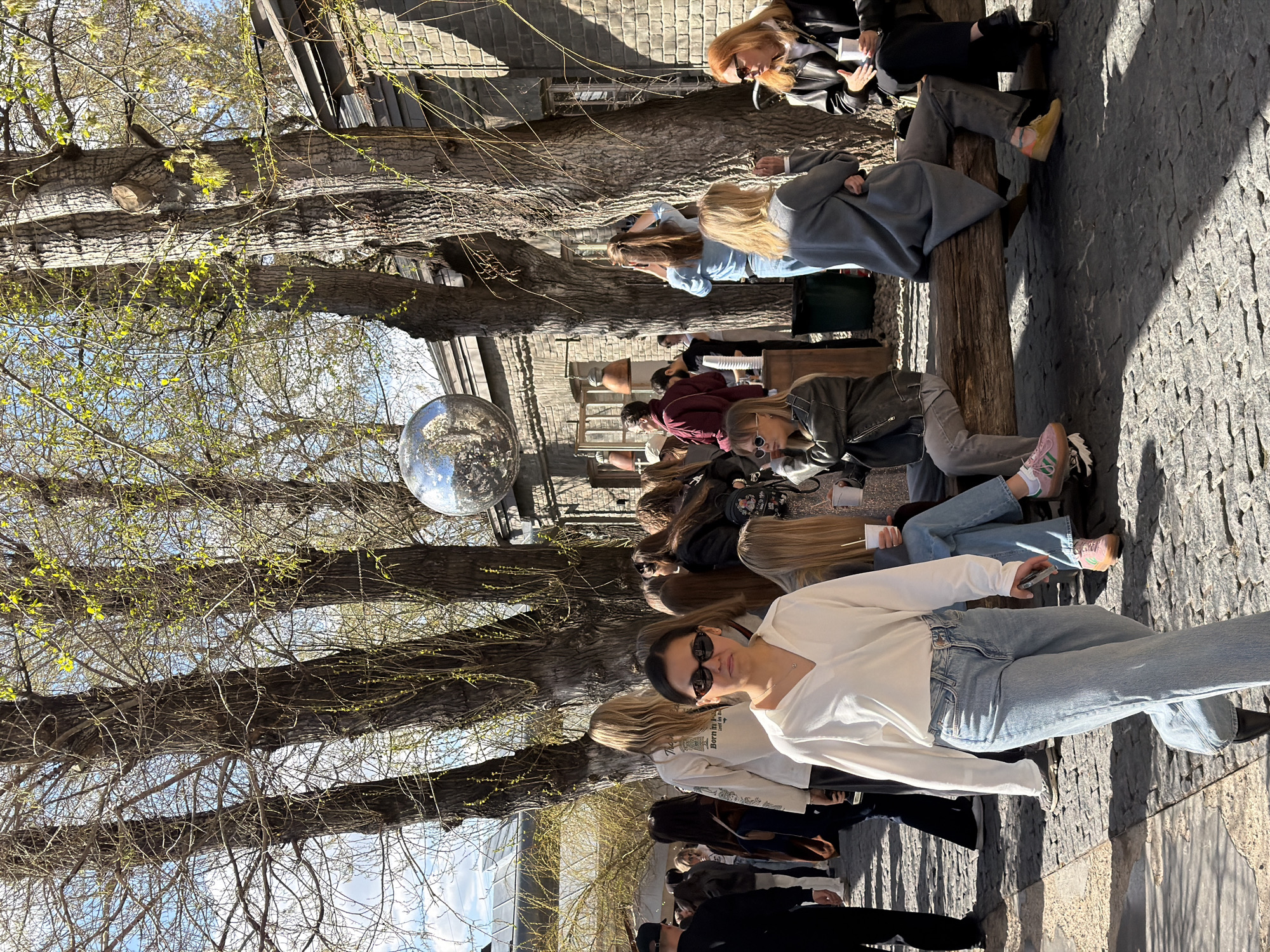 A row of people sit along a wooden beam, with trees overhead and a silver disco ball hanging among the branches. The seated individuals wear casual clothing. The flooring appears to be paved with uneven stones. This scene is likely located in Gorky Park in Moscow, Russia.