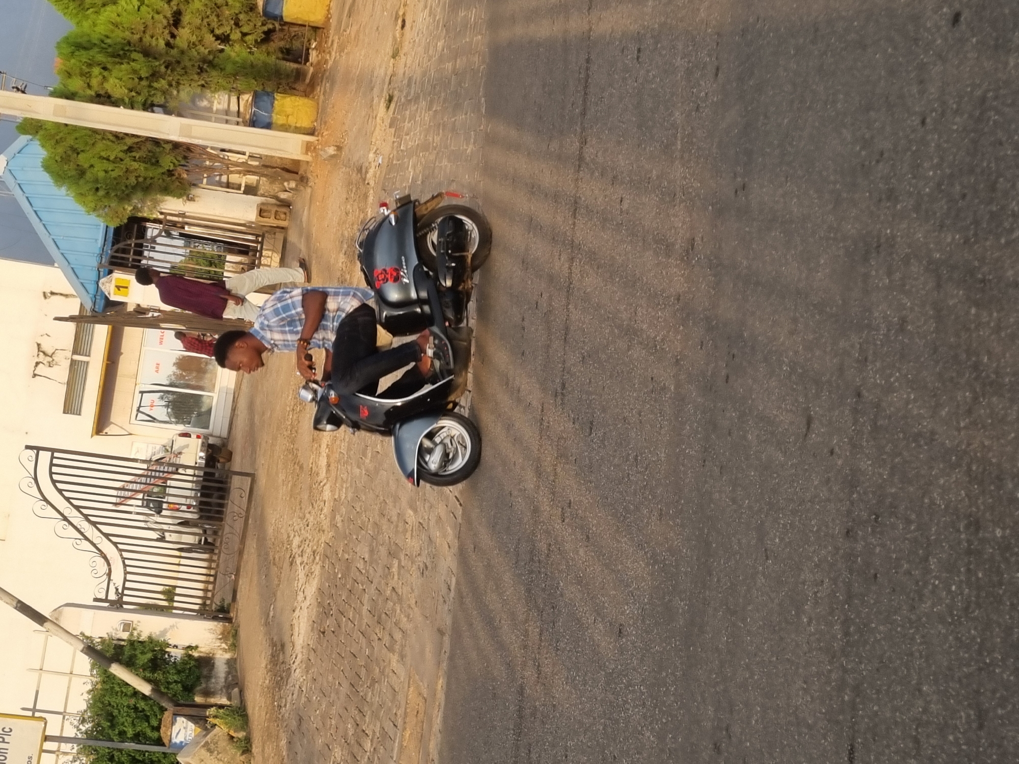 The image captures a scene in Jos, Nigeria, featuring a man dressed in a blue plaid shirt and black pants astride a dark-colored scooter. He is positioned on a paved area that transitions into a road with visible cracks and markings. To the left, there's a building with a light-colored exterior, a blue awning, and a decorative iron gate. Adjacent to the building is a patch of greenery. In the background, another individual is walking away from the building. The overall lighting suggests it is daytime.