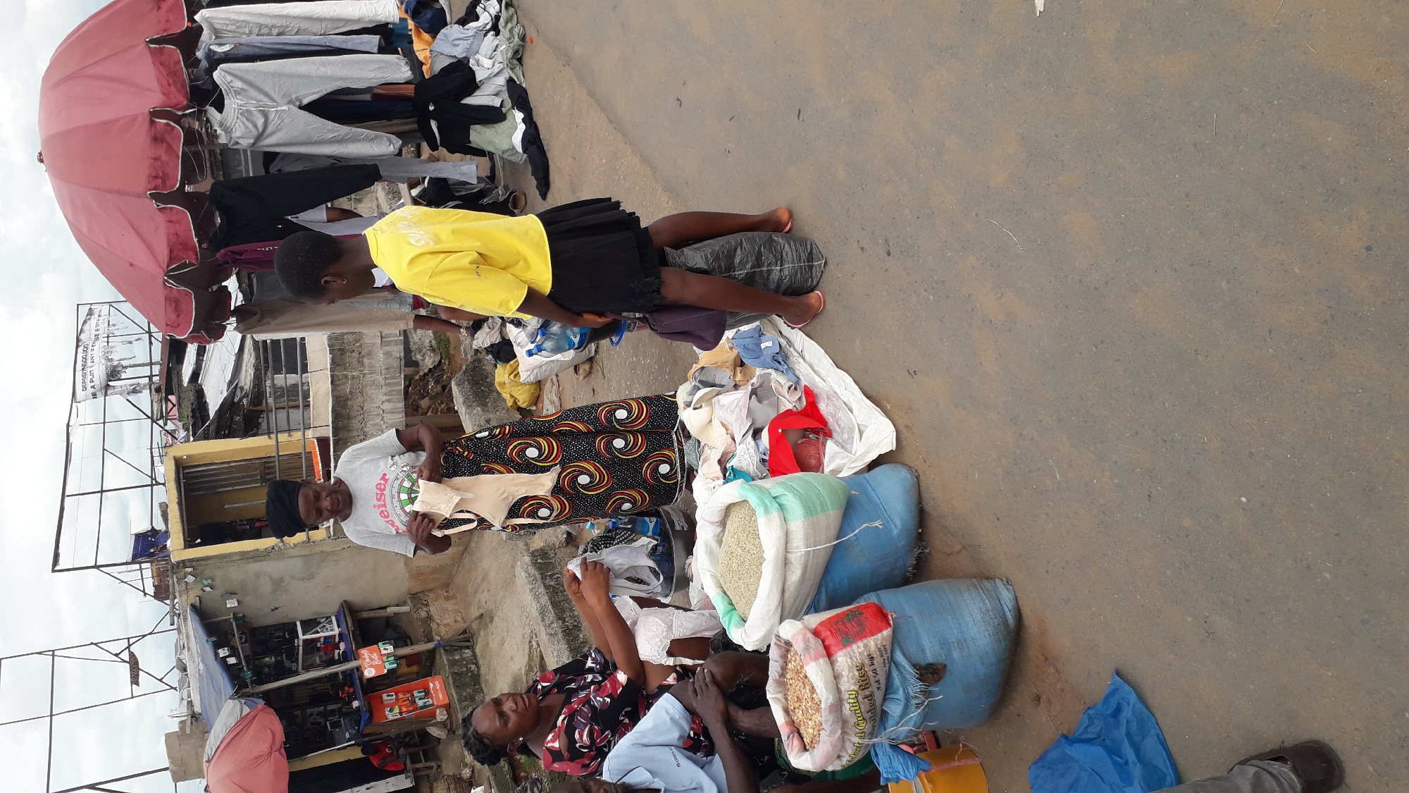 This image captures a bustling outdoor market scene in Onitsha, Nigeria. The primary subjects are several people, appearing to be vendors and shoppers, interacting amongst a diverse array of goods.
The scene is dominated by the display of clothing, with items hanging on racks under a large, open pink umbrella, suggesting a market stall. Piles of clothing are also scattered on the ground, interspersed with sacks of grains and other produce. One sack is clearly labeled with "Colort & Chality" and "led Rice" with "Nel 50 kgs." Another sack of grain, possibly corn, is also visible.
A young woman in a bright yellow t-shirt and black skirt is seated on a large dark-colored sack, looking towards the ground. Another woman, wearing a t-shirt with the word "weiser" printed on it, is holding up a light-colored undergarment, possibly for display or sale. A third woman in a patterned dress and a headscarf is also present, along with other individuals whose faces are partially obscured.
The setting appears to be a somewhat rough street or market ground, with a mix of concrete and dirt. In the background, there are buildings and structures, typical of an urban market area. A sign is partially visible with text that includes "DEPOSITN500.000" and "RE A PLOT ANY IFTIGE E". The sky appears overcast, suggesting a cloudy or potentially humid day, common in this region. The overall impression is one of active commerce and everyday life in a Nigerian market.