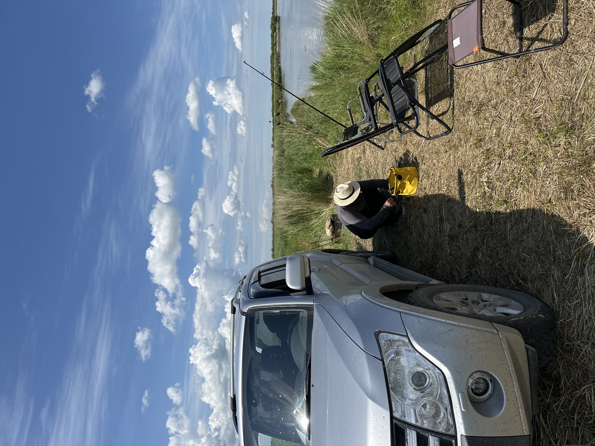An outdoor, eye-level, medium shot captures a silver SUV parked on a dirt path next to a body of water on a sunny day. A person wearing a wide-brimmed hat is crouched beside a yellow bucket, seemingly engaged in an activity near the vehicle. Two black folding chairs and a small brown stool are set up on the grass, implying a resting or waiting area. A fishing rod stands upright by the water's edge. The landscape is characterized by tall, dry grass and lush green reeds lining the water, with a clear blue sky dotted with fluffy white clouds overhead. The scene depicts a tranquil, possibly recreational outing in a natural setting. The image does not provide specific details or landmarks that would allow for precise location identification within the city of Im. Tel'mana, Russia.