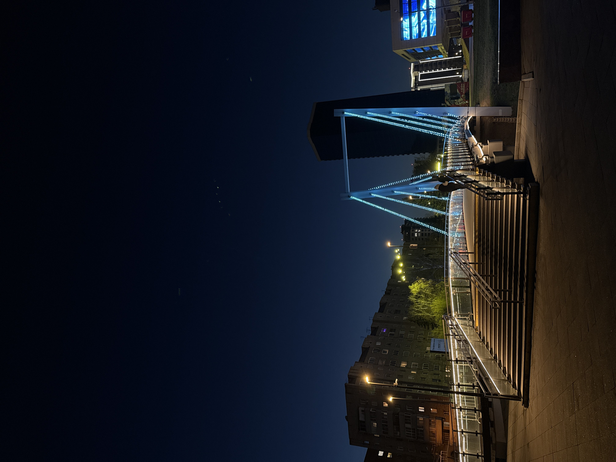 The image depicts a night scene in Krasnoyarsk, Russia. A pedestrian bridge with blue lights spans the foreground, leading up from a set of stairs with stainless steel handrails. A modern building with illuminated blue windows stands to the right of the bridge, on the other side. Residential buildings with illuminated windows and streetlights line the horizon behind the bridge. The sky is a dark shade of blue. The perspective is a low angle, emphasizing the height of the bridge and the buildings. The ground is paved with stone.