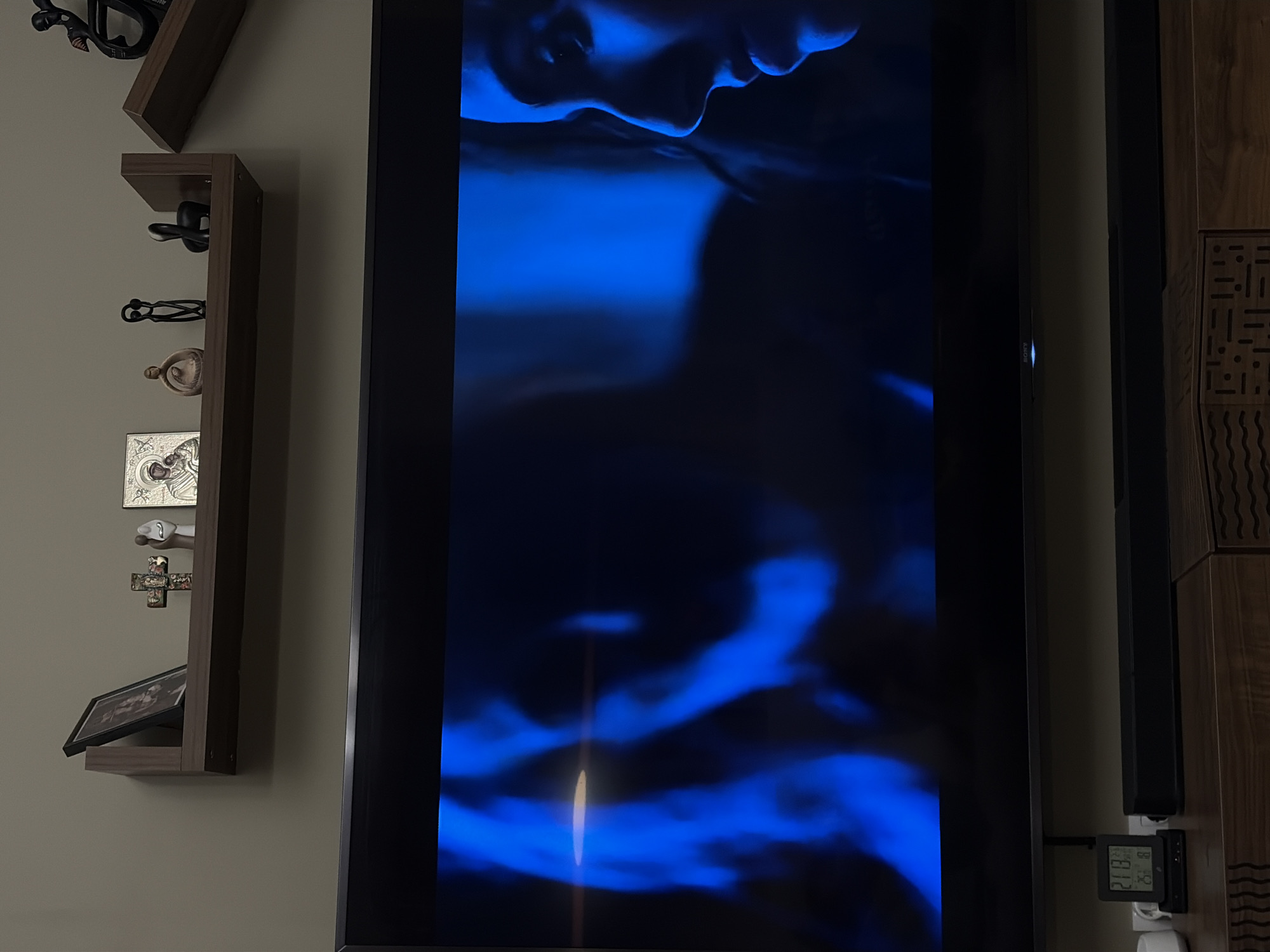 An indoor scene captured in dim lighting showcases a large, modern flat-screen television displaying a vivid blue image. The screen occupies the central and right portion of the frame, showing the profile of a person's face, possibly submerged in water or dramatically lit with blue light, creating a mysterious and artistic effect. A subtle "SONY" logo is visible on the lower bezel of the television.
To the left of the television, a vertical, multi-tiered wooden shelf unit is mounted on a light beige wall. The shelves hold various decorative items: several dark, abstract sculptural pieces, a light-colored figurine, a silver-toned religious icon (possibly depicting the Virgin Mary and Christ Child), a small, ornate cross, and a framed picture propped up on one of the lower shelves.
To the far right of the television, a section of wooden wall paneling or furniture is visible, featuring intricate carved or patterned designs. A black, rectangular object, possibly a soundbar or speaker, is mounted vertically between the television and the wooden panel. Below this, a small digital display unit shows "21.8" (likely temperature), "82" (possibly humidity), and a time reading that appears to be "20:13". The ambient lighting suggests it is evening or night in Sofia, Bulgaria. The overall impression is a cozy, modern living space.