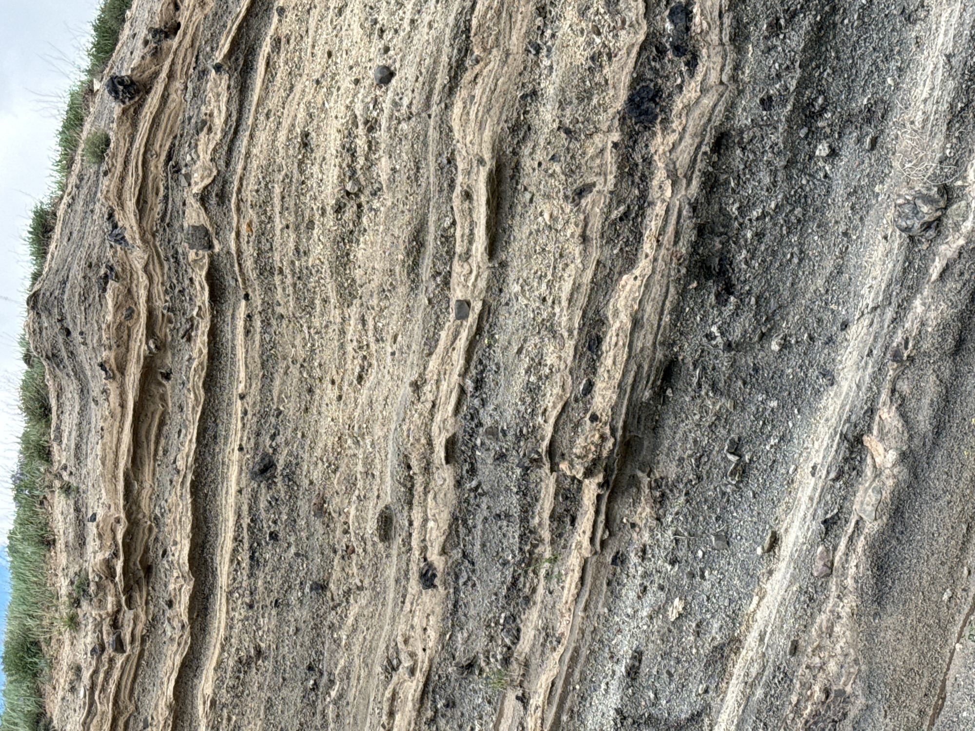 The image displays a close-up view of a rock formation, characterized by distinct horizontal layering. The strata vary in color, with bands of light tan, gray, and darker shades of gray and black. These layers are composed of smaller rock fragments, pebbles, and volcanic ash, suggesting a history of deposition and erosion. Some larger, dark-colored rocks are embedded within the layers. Sparse green vegetation, appearing as grass and small bushes, grows along the top of the formation. The sky is visible in the upper background, appearing overcast with a pale blue hue peeking through. The scene is static, with no visible people or animals. The environment appears to be an outdoor geological outcrop. Based on the provided information, this rock formation is located in the city of Tsghuk, Armenia.