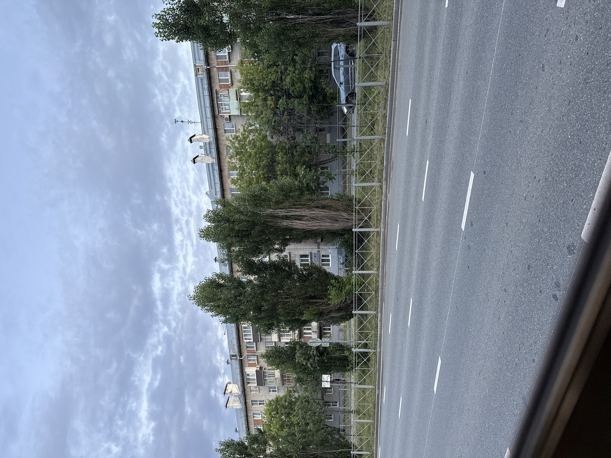 The image depicts a street scene in Kazan, Russia. In the foreground, a wide asphalt road is visible with white lane markings. A metal guardrail separates the road from a grassy median and a sidewalk. Beyond the sidewalk, a multi-story apartment building with a brick facade and numerous windows stands. Trees and other vegetation are interspersed along the front of the building and along the street. A dark-colored car is driving on the road, partially obscured by trees. The sky is overcast with gray clouds. The overall impression is an urban residential area on a cloudy day.