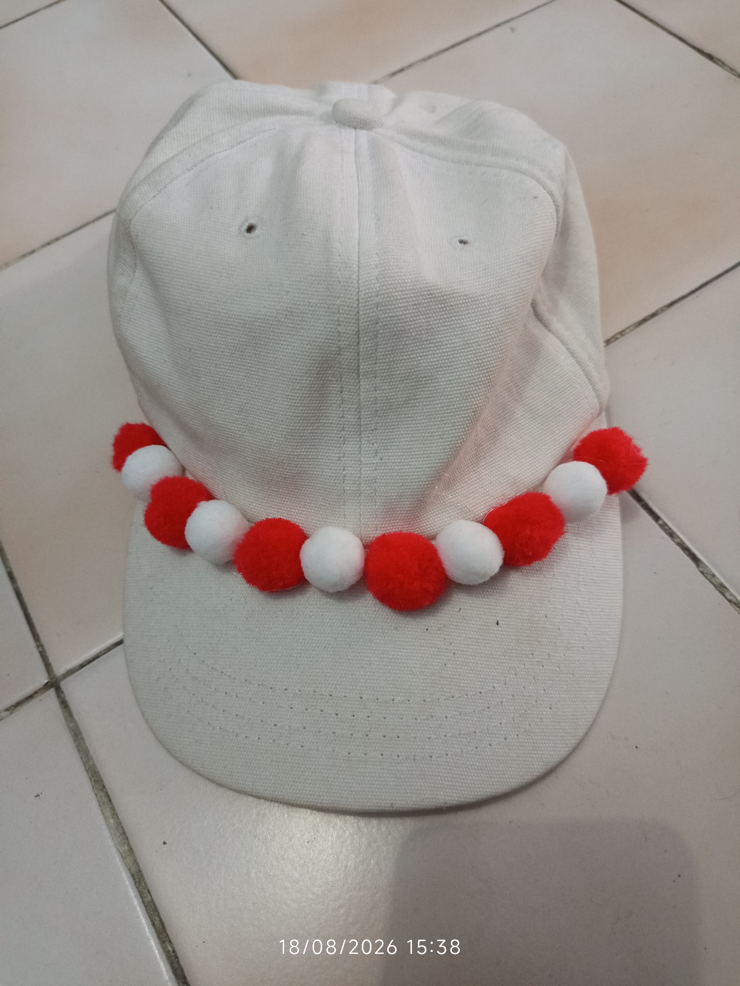 A white baseball cap is placed on a tiled floor. A string of red and white pom-poms is wrapped around the base of the cap, near the brim. The tiles are cream-colored and laid in a grid pattern. The timestamp "18/08/2026 15:38" is visible in the lower right corner of the image. The scene appears to be indoors, likely in a home or personal space, and does not provide specific details about Canggu, Indonesia.