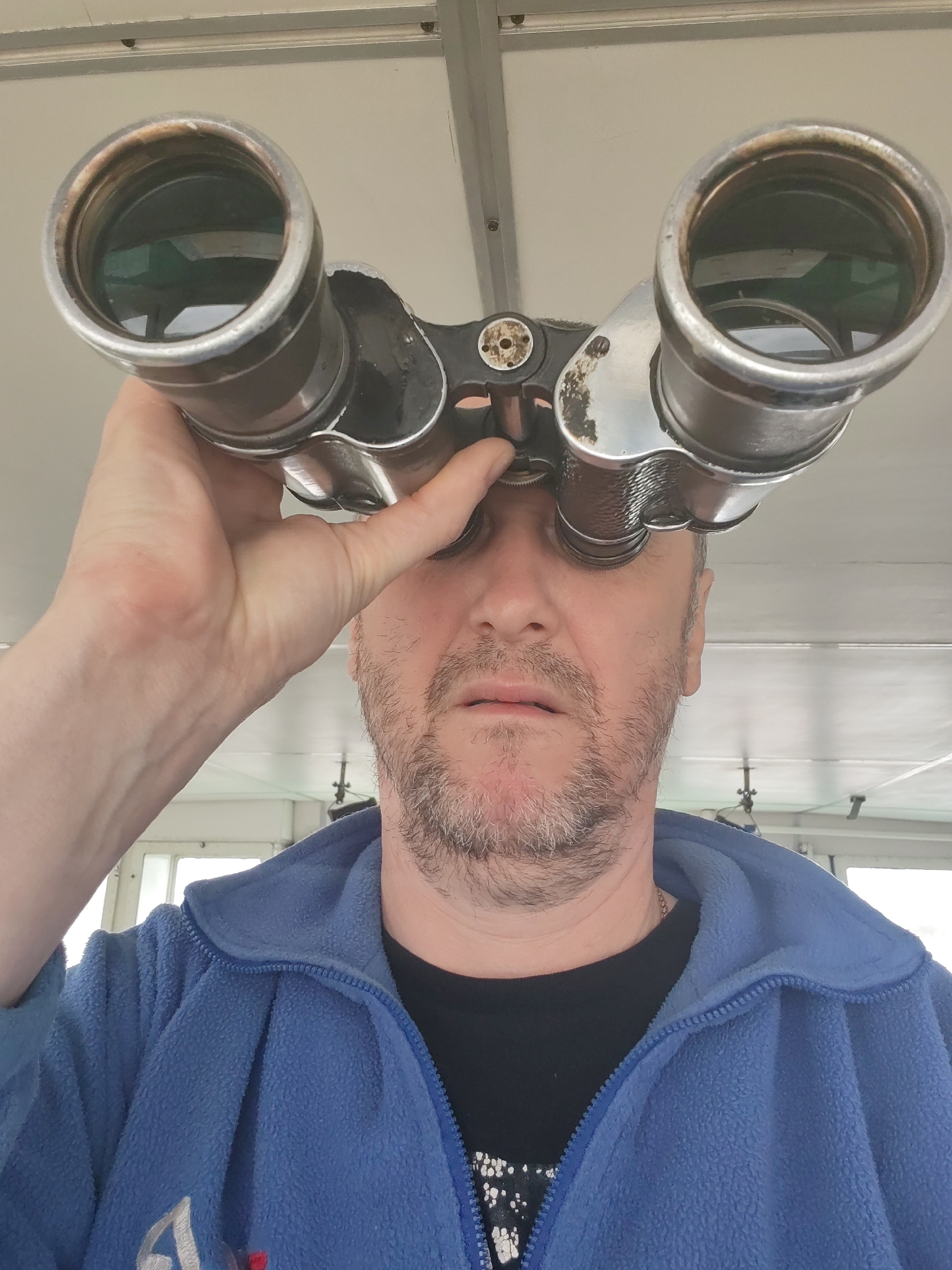 A man with a beard is looking through binoculars. He is wearing a blue fleece jacket over a black t-shirt. The binoculars appear to be vintage or antique. The setting seems to be indoors, possibly on a ship or a vessel, given the overhead structure and windows. The city or location is not discernible from the image.