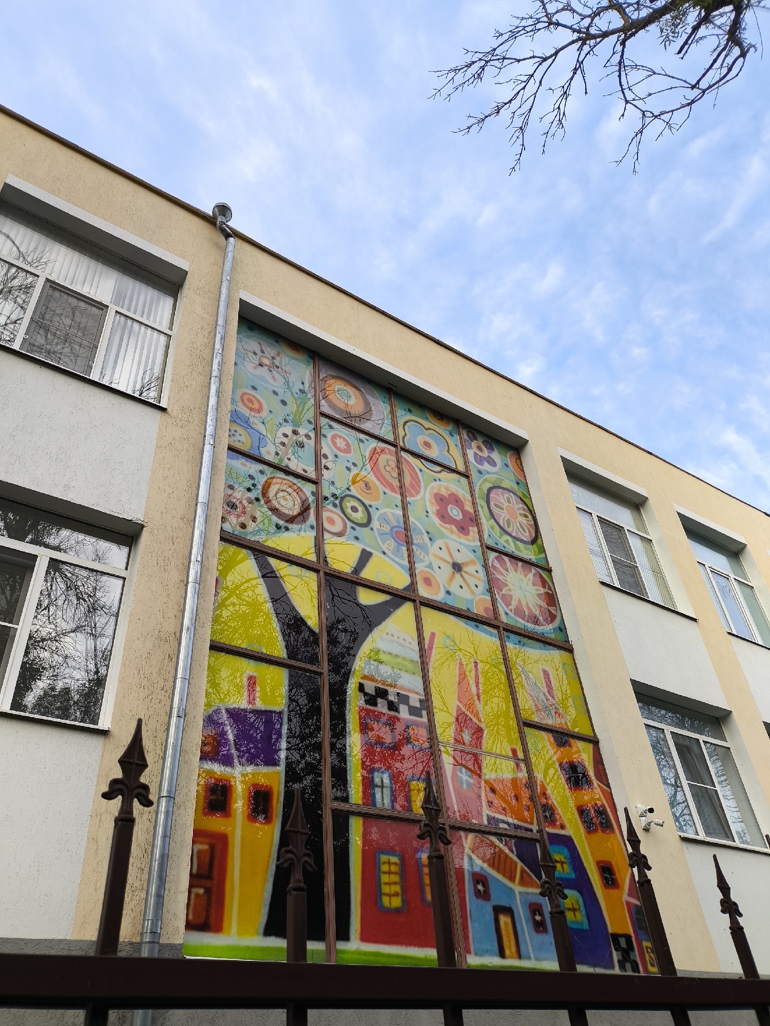 The image shows the facade of a building with a large, colorful stained-glass window. The window depicts a stylized village scene with houses and a tree, rendered in bright, abstract shapes and patterns. The building itself is light beige with white window frames and appears to be multi-story. A metal downspout runs along the side of the building next to the stained-glass window. In the foreground, there is a dark metal fence with decorative finials. The sky above is blue with scattered white clouds, and the bare branches of a tree are visible against the sky. The scene is captured from a low angle, looking up towards the building. The location is identified as Yessentuki, Russia.