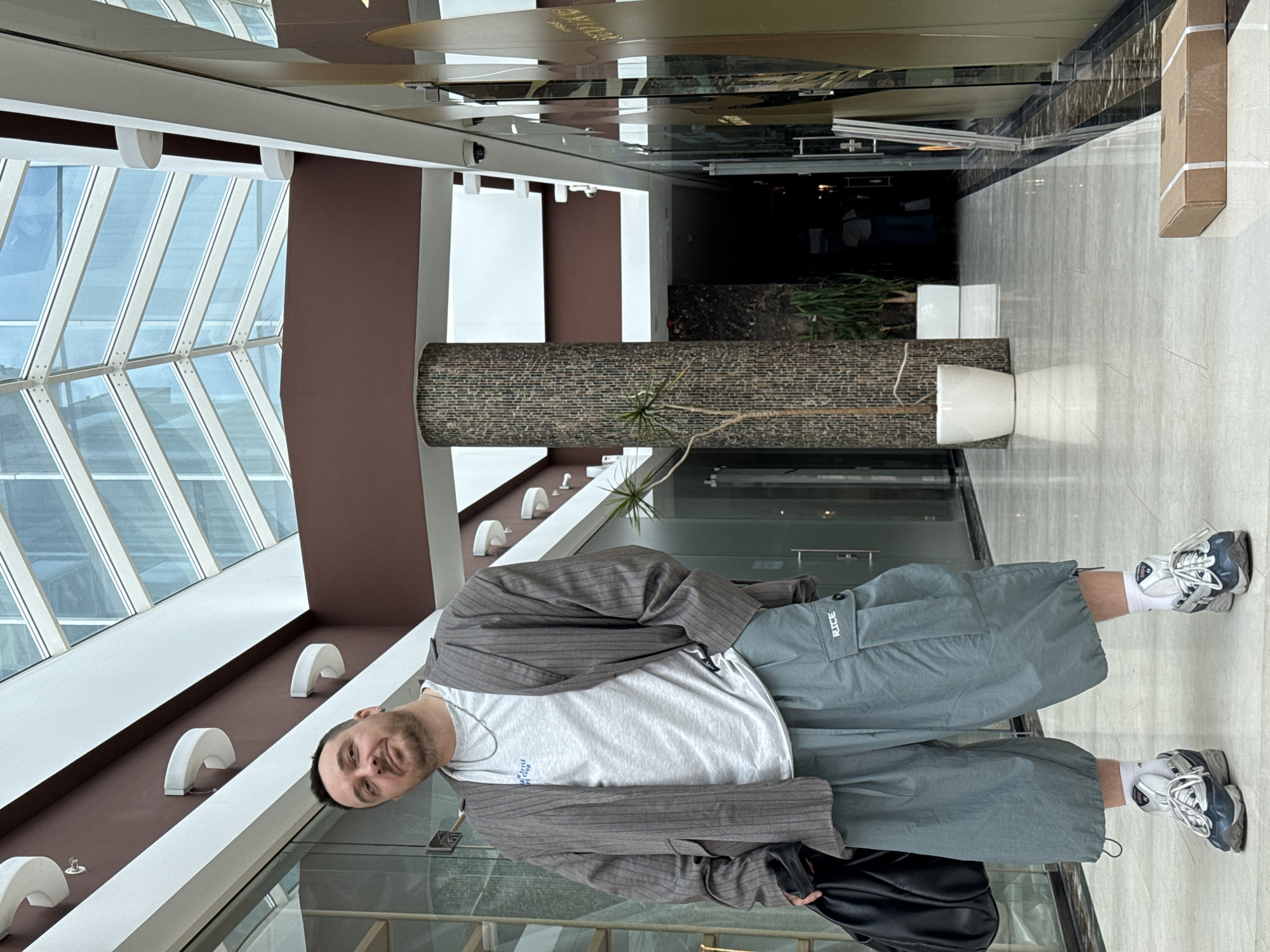 A man is standing in the glass-walled atrium of a building, apparently in Budva, Montenegro, based on the location metadata. He is wearing a grey pinstripe jacket, white t-shirt, light grey cargo shorts, white socks, and grey and blue sneakers. He is also holding a black bag in his left hand. The atrium has a high, angled glass ceiling and brown walls with embedded, half-moon shaped light fixtures. A long, cylindrical planter filled with plants runs along the side of the atrium. The floor is light-colored tile.