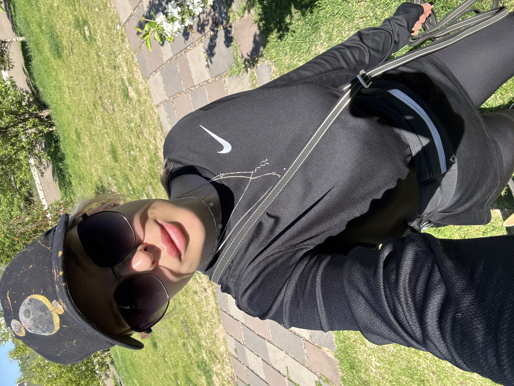 The image features a person outdoors on a bright, sunny day, likely in a park or garden setting in Krasnoyarsk, Russia. The shot is a self-portrait, showing the individual from a slightly high angle.
The subject is wearing a black long-sleeved athletic top with a white Nike swoosh logo on the chest, paired with black leggings. A black cap, decorated with a distinct yellow and gray design resembling a moon or other celestial body, is worn on their head. They are also wearing large, dark sunglasses. A delicate, silver-colored necklace is visible around their neck, and a black fanny pack with a gray stripe is secured around their waist. A thin, light-colored strap crosses their chest diagonally, possibly belonging to the fanny pack or another small bag. In their right hand, they hold several black and white striped straps, which appear to be leashes. The person's lips are slightly parted, suggesting a subtle smile, and strong sunlight casts shadows across their face.
The background reveals a paved path made of alternating light and dark gray rectangular stones, adjacent to vibrant green grass. Green trees and bushes are visible on the periphery, and a branch with white blossoms extends into the frame from the upper right, casting shadows on the path. The bright illumination and distinct shadows indicate a clear, sunny day. The overall impression is one of relaxation and enjoyment of the outdoors.
The only visible text is the "Nike" logo on the subject's shirt. The cap also features a prominent pictorial design of a moon.
