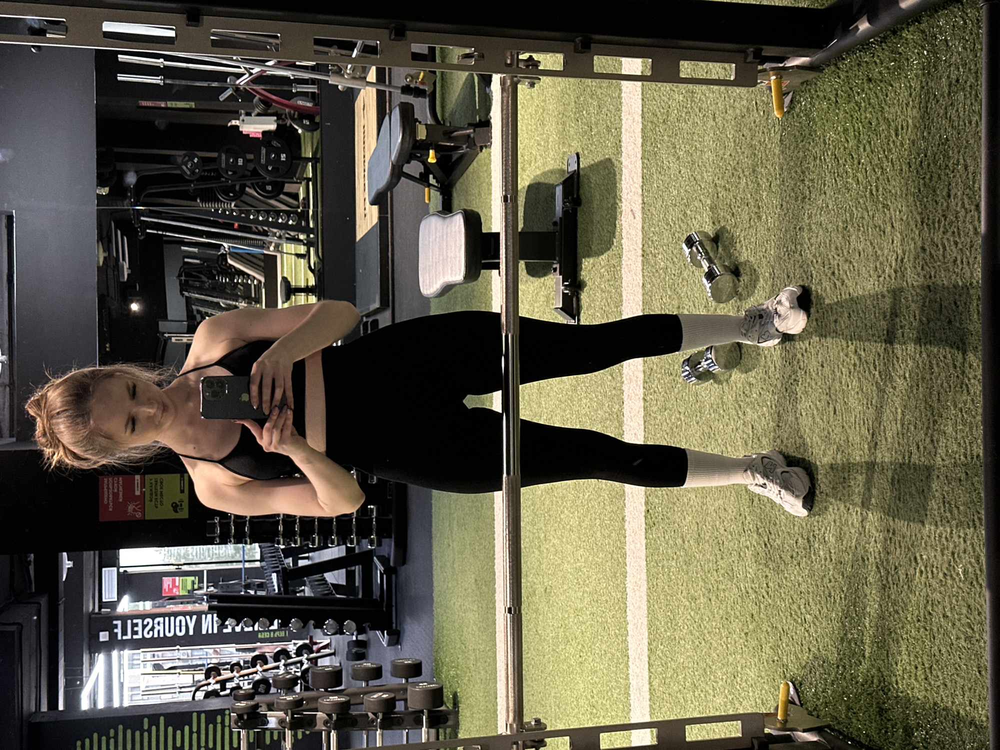 A young adult female is depicted in a full-body mirror selfie taken within a gym located in Unknown, Unknown. She is wearing a black sports bra, black high-waisted leggings, white crew socks, and light-colored New Balance athletic shoes. Her light-colored hair is pulled up into a messy bun. She holds a dark-colored smartphone with both hands, looking down at the screen.
The foreground is framed by components of a dark-colored squat rack or Smith machine, with a metallic barbell positioned horizontally at waist height. Two chrome dumbbells rest on the green artificial turf floor to the right of the subject's feet. The turf features a white horizontal dividing line across the mid-ground. Behind the subject, two workout benches, one gray and one black, are visible on the turf.
The background primarily consists of a large mirror reflecting various gym equipment. On the left side of the reflection, a rack of hexagonal dumbbells, a wall with vertical green striping, and an inverted sign displaying Cyrillic text including "УДОБНОЕ", "МОЩНОЕ", and "БЫСТРОЕ" are present. The right side of the reflection shows multiple weight machines, barbells, and stacked weight plates, some visibly marked with "10" and "15". The gym environment features dark walls and artificial overhead lighting, which casts shadows from the subject and equipment onto the turf.