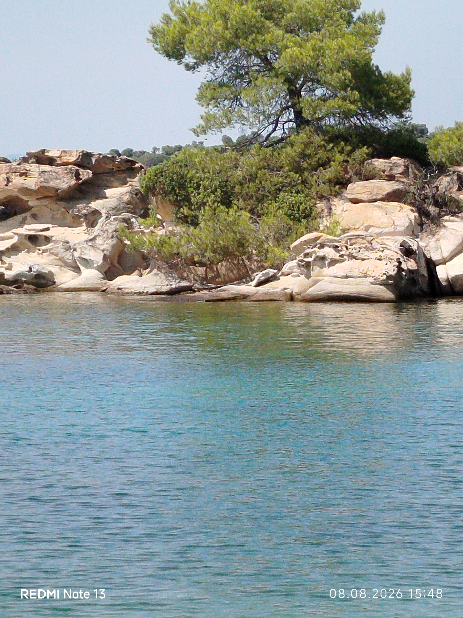 The image depicts a serene coastal scene in Sithonia Municipal Unit, Greece. The foreground is dominated by calm, turquoise water with gentle ripples reflecting the sunlight. In the middle ground, a rocky shoreline is visible, characterized by weathered, light-colored stones and outcroppings. Lush green vegetation, including trees and shrubs, grows on top of and around the rocks, with a prominent pine tree with dense foliage at the center-right of the frame. The sky above is a clear, pale blue. The bottom right corner of the image displays a timestamp reading "08.08.2026 15:48" and the text "REDMI Note 13" is present in the bottom left corner. There are no people or animals visible in the image.